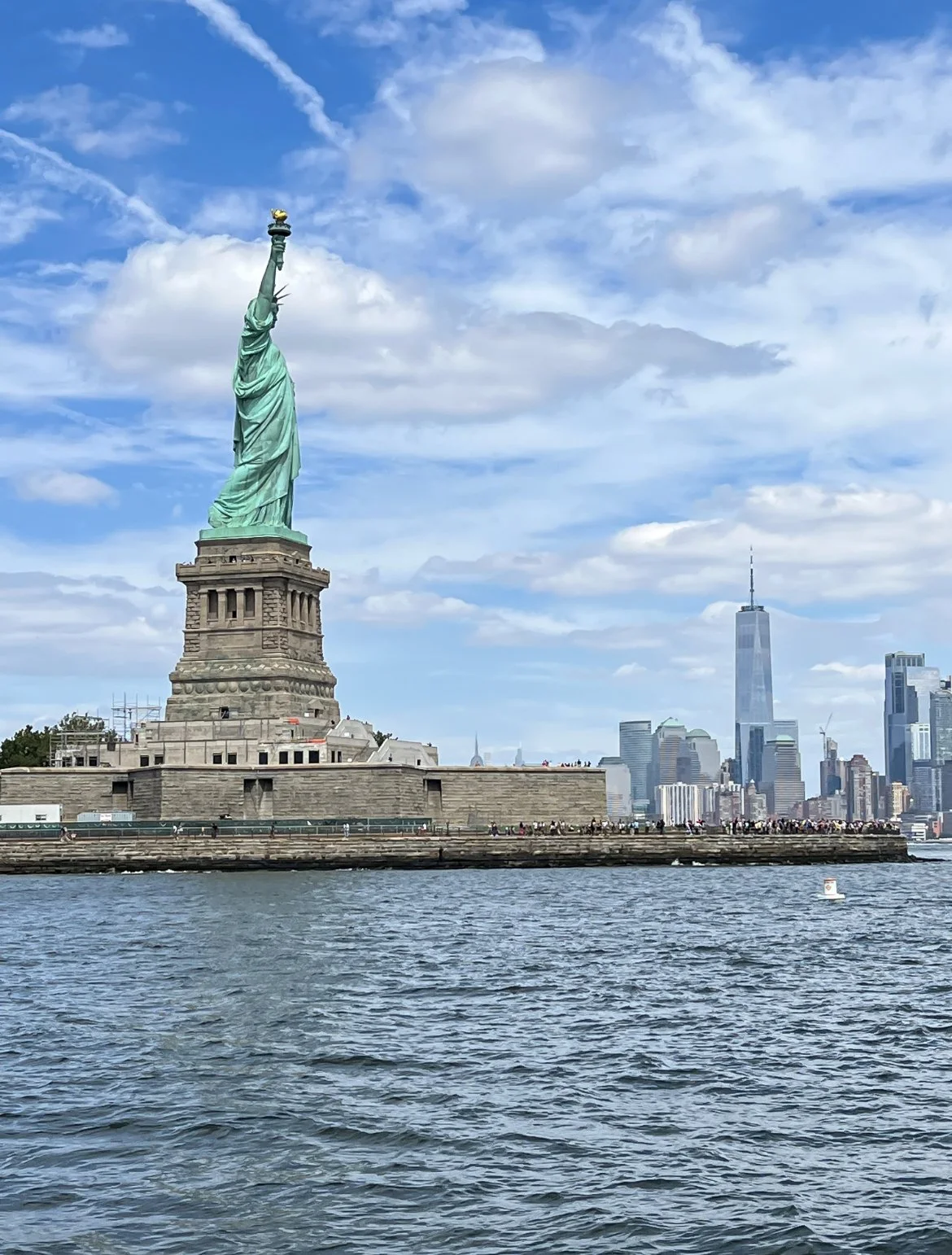 The Statue of Liberty on Liberty Island in New York Harbor with a city skyline including One World Trade Center in the background under a partly cloudy sky.