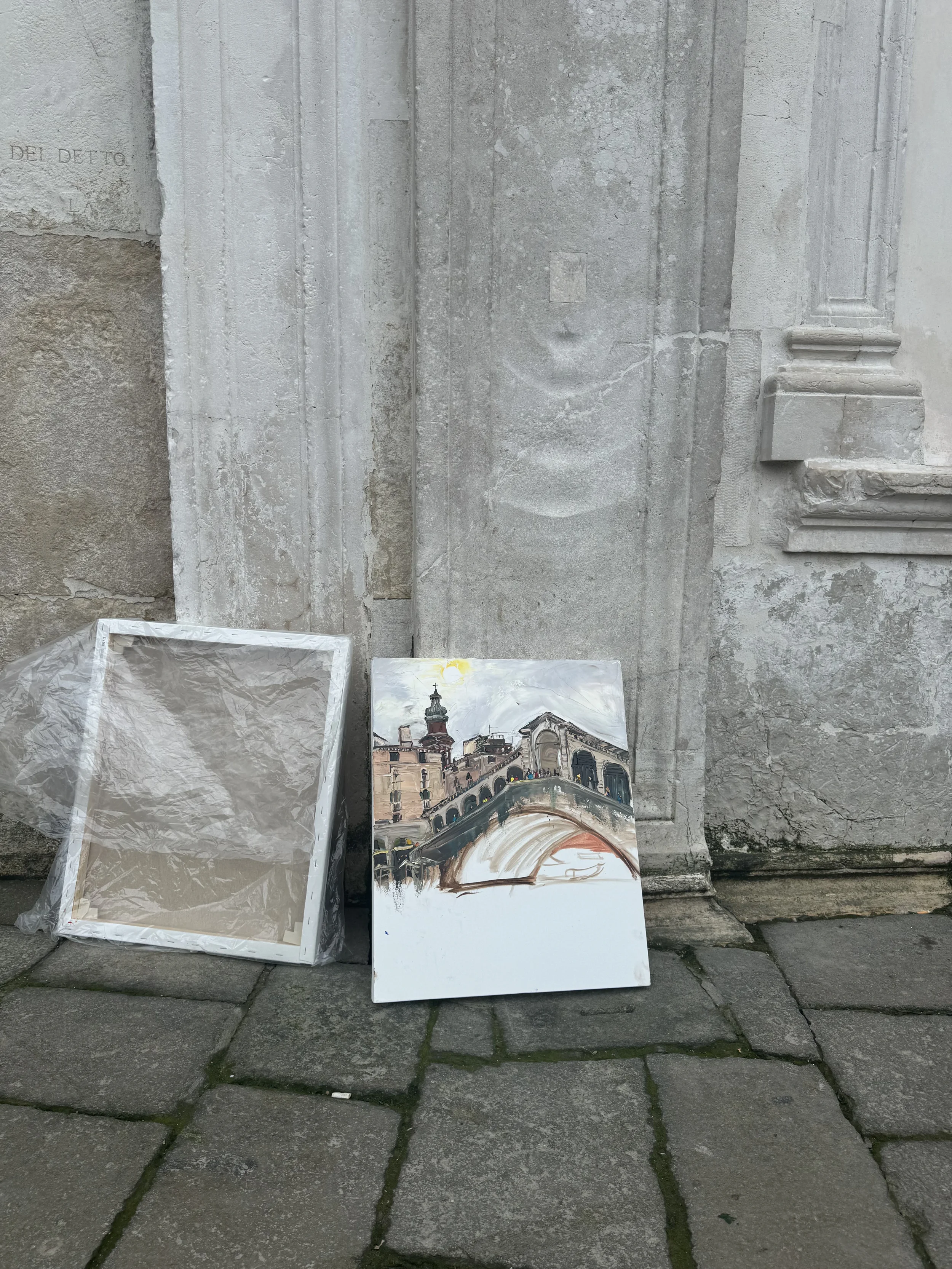 A painting of a historic urban scene with a bridge, buildings, and a church with a tall steeple. The painting is leaning against a stone wall on a cobblestone street.