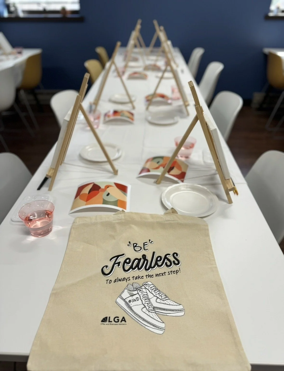 A long table set for a group activity with small easels, printed sheets with a colorful abstract design, white plates, and pink water glasses. A beige tote bag with the words 'Be Fearless' and a drawing of sneakers is in the foreground, and the backg