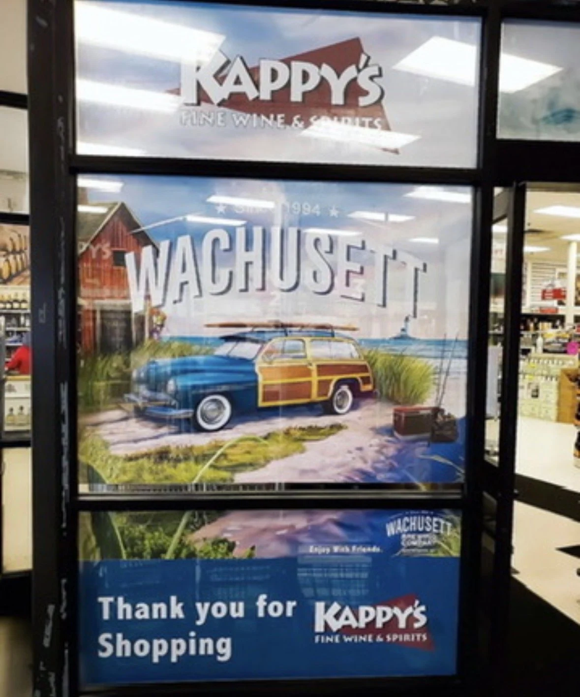 Store window display for Kappy's Fine Wine & Spirits with a colorful illustration of a vintage car parked on a beach with grass and a wooden shed in the background, and a sign thanking customers for shopping.