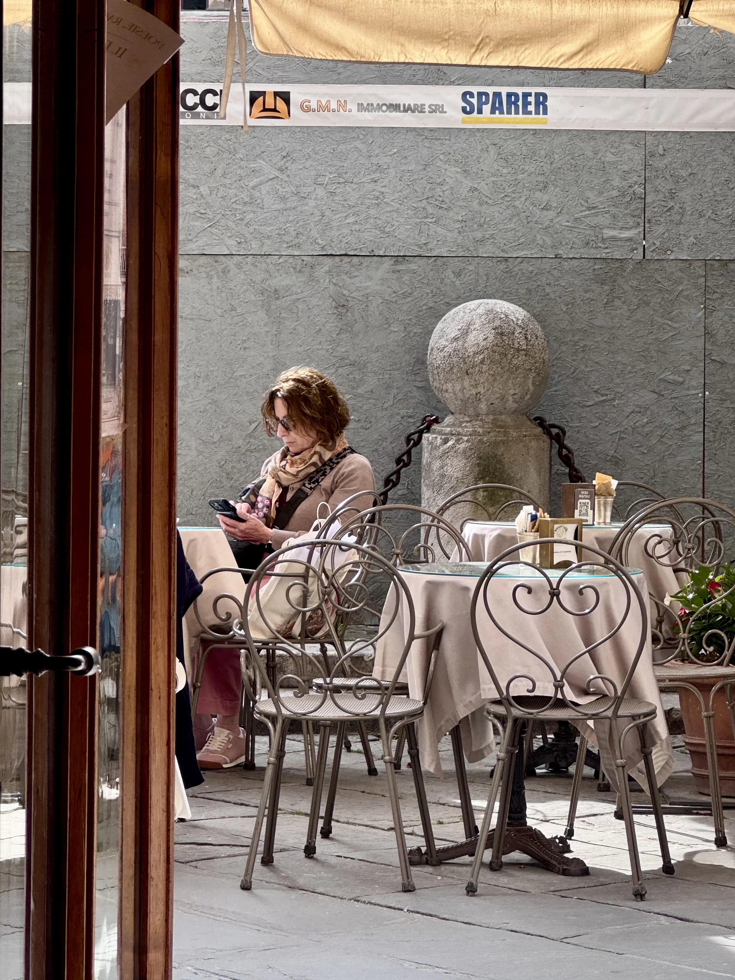 A woman with brown curly hair, sunglasses, and a patterned scarf sits at a table in an outdoor cafe, looking at her phone. The cafe has empty chairs with ornate metal backs and white tablecloths. A large stone sphere on a pedestal and a textured conc