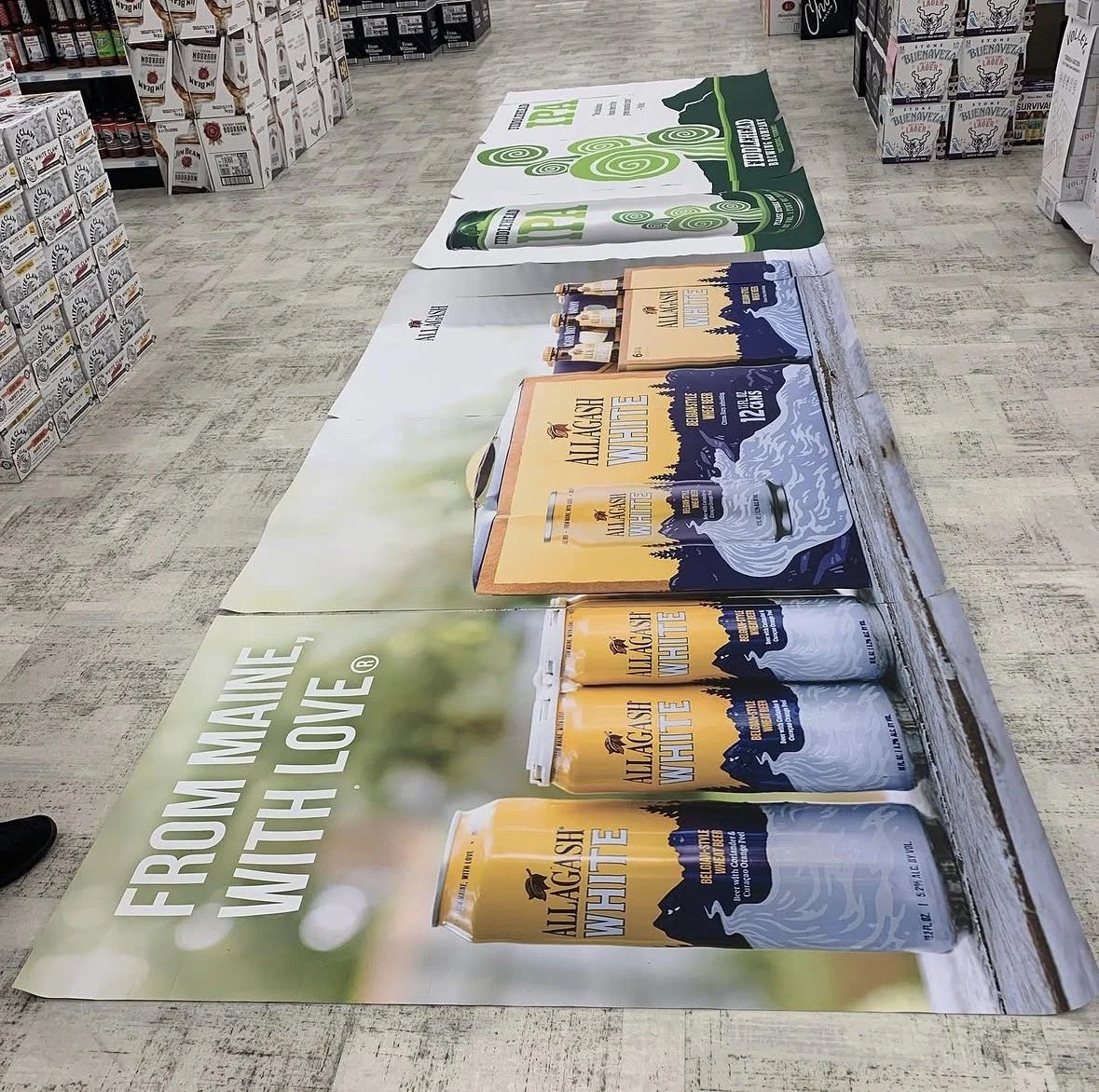 A promotional display in a store advertising Allagash White beer with large images of beer cans and bottles, along with a background of a green forest and mountains.