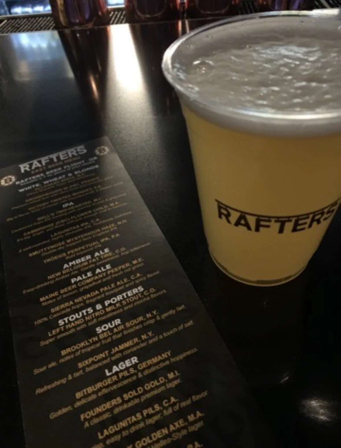 A pint of beer with a yellow sleeve labeled 'RAFFTERS' next to a printed beer menu on a black table.
