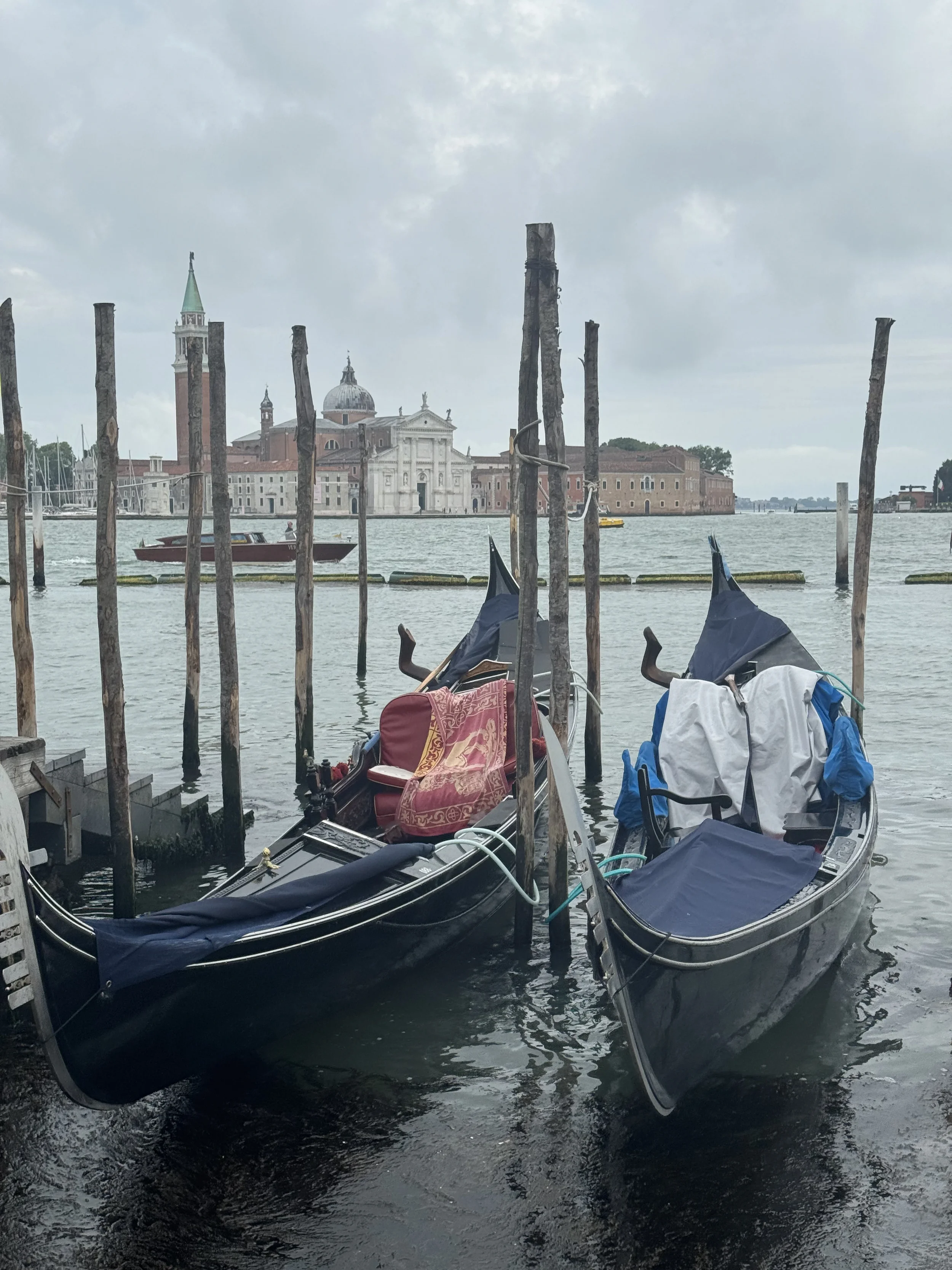 Two gondolas docked at a pier with weathered wooden posts, set against a backdrop of historic buildings in Venice, Italy, on a cloudy day.