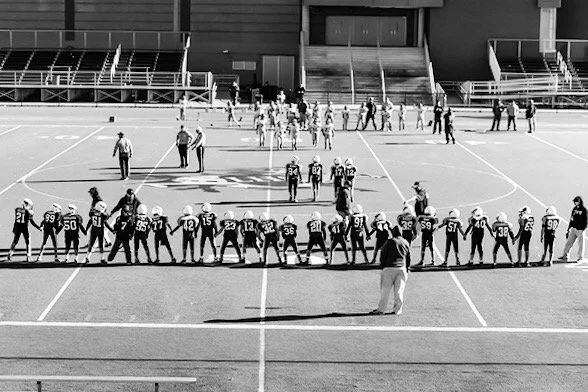 High school football players in helmets and jerseys lined up on the field with coaches and officials during a game or practice.