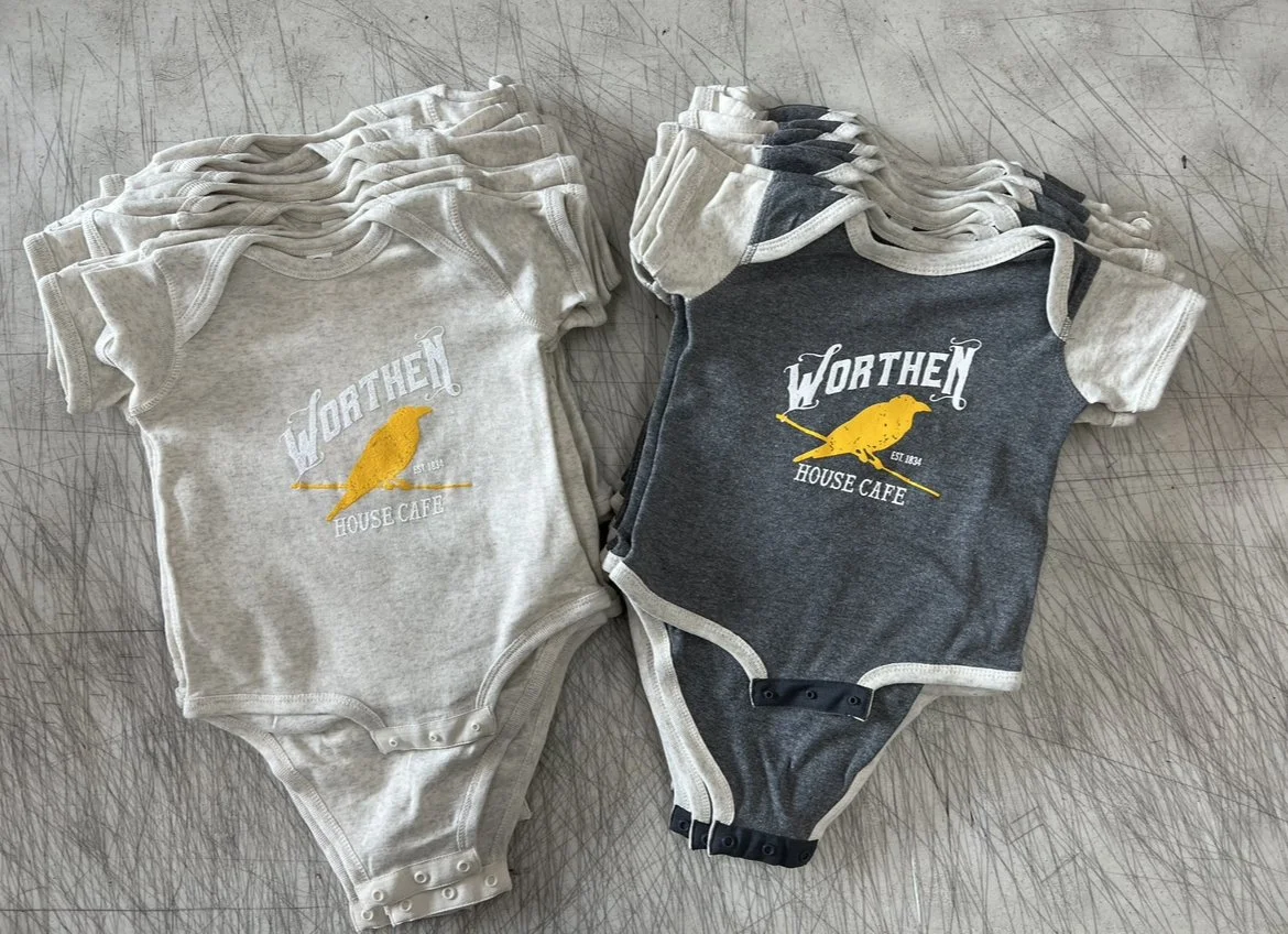 Stacks of baby onesies, some in light gray with Worthern House Cafe logo, and others in dark gray with the same logo, on a surface with scratch marks.