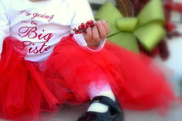 Child wearing a white shirt with red text and a red tutu, holding a small bunch of red berries, sitting next to a large green bow.