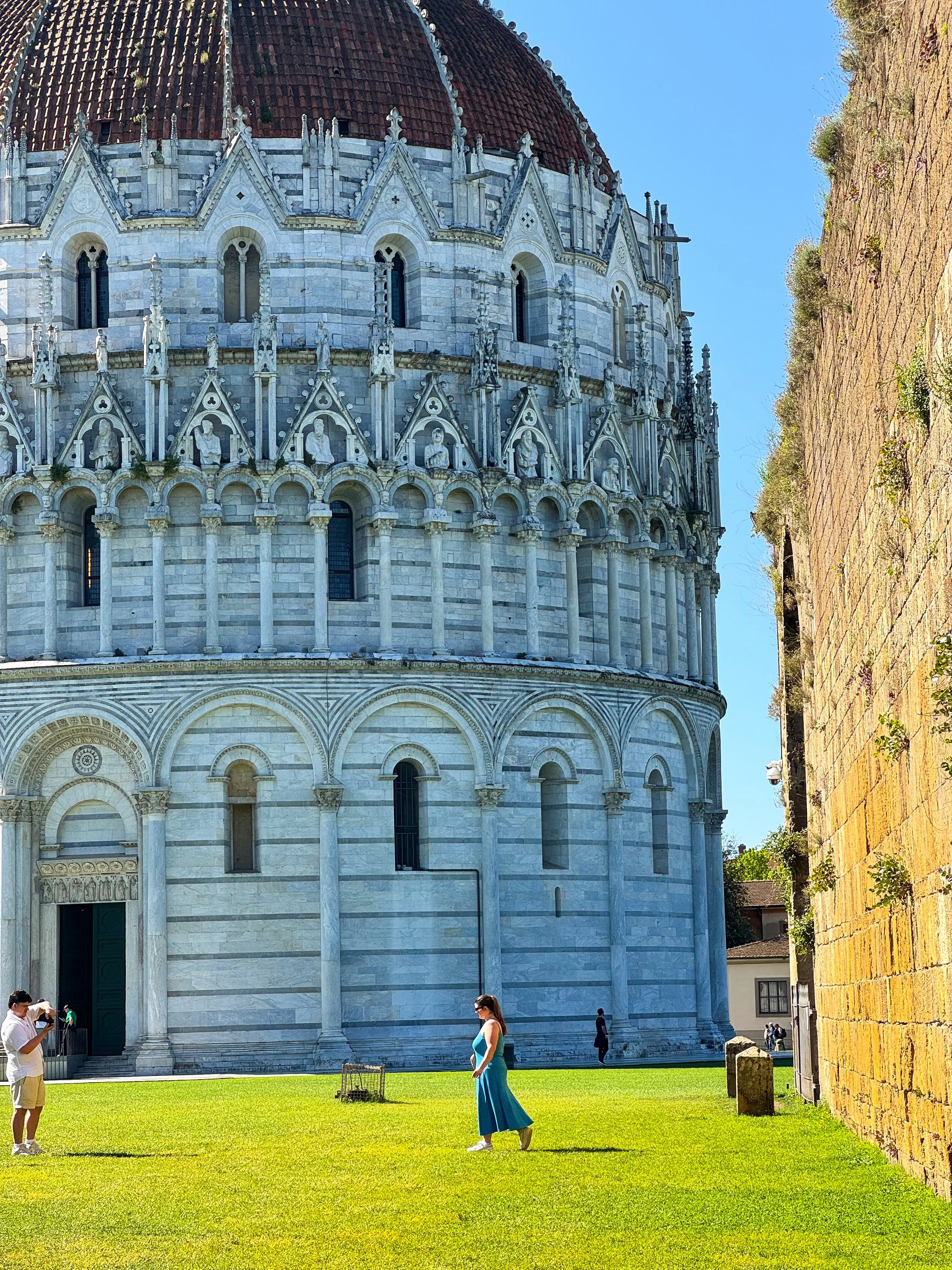 People standing and walking on the grass in front of a historic building with ornate stone architecture and statues, under a blue sky.