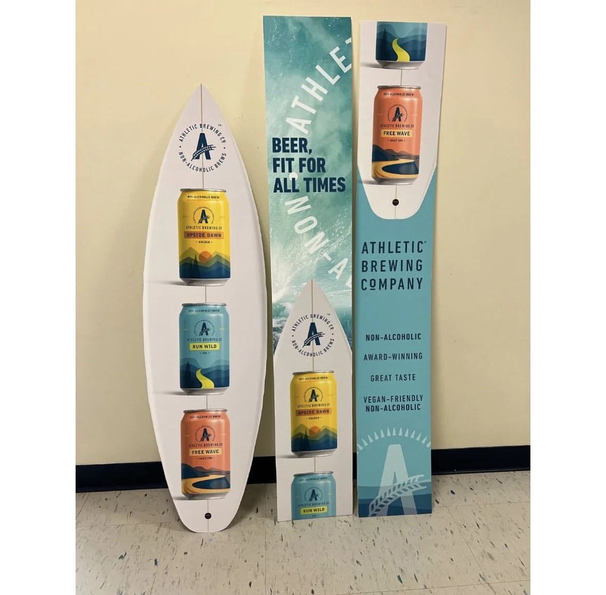 Two promotional stands with Athletic Brewing Co non-alcoholic beers. The left stand features three cans labeled 'Upside Dawn,' 'Run Wild,' and 'Free Wave,' with a surfboard shape. The right stand displays a single can 'Free Wave' on a beach-themed ba