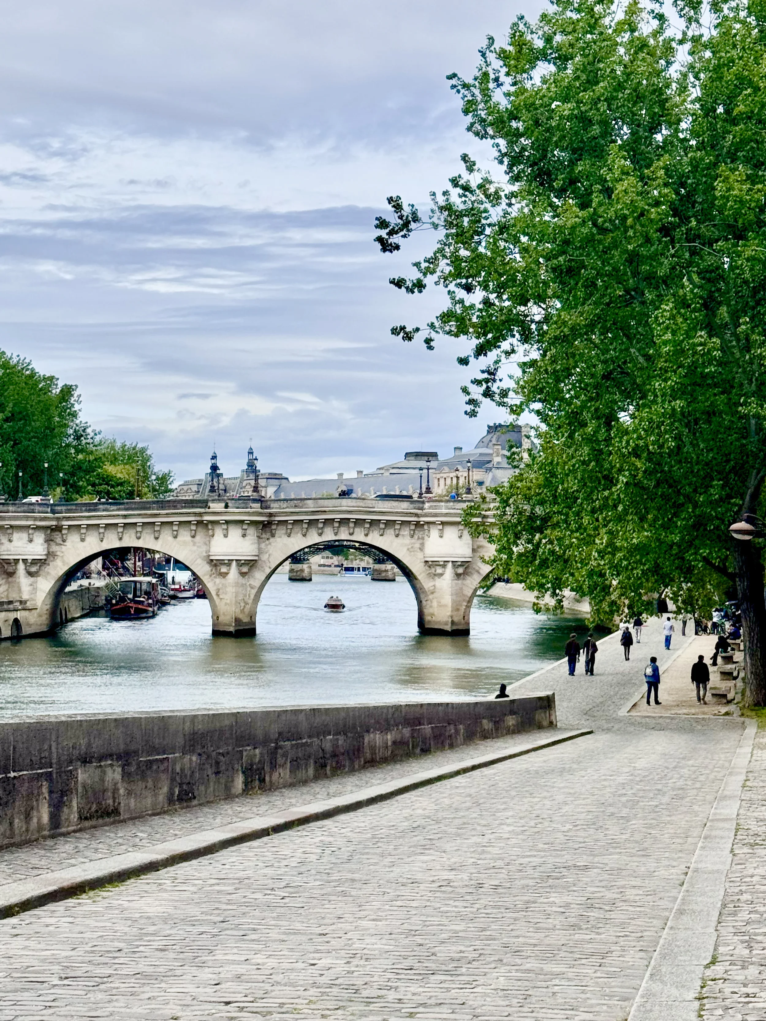A stone bridge over a river with boats, and a paved walkway with people walking beside it, trees on one side, under a cloudy sky.