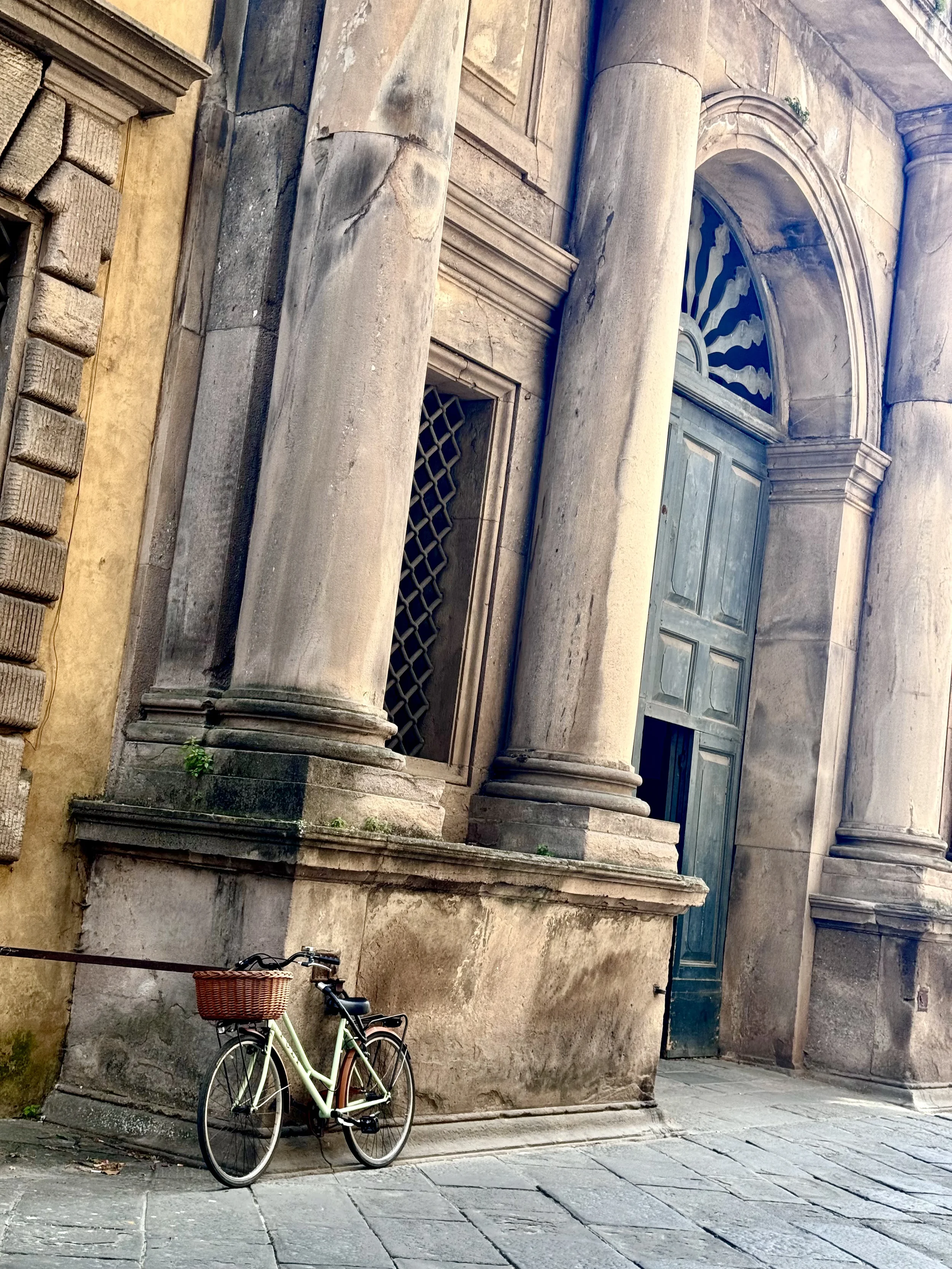 A pale green bicycle with a wicker basket parked against a weathered stone and stucco building wall on a cobblestone street.