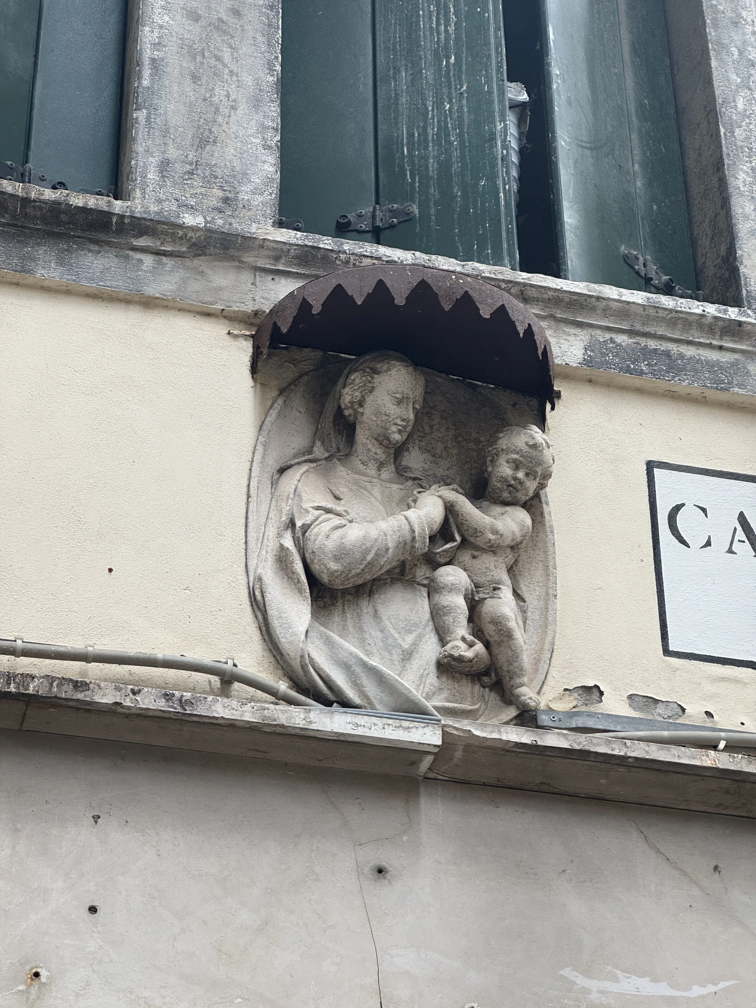 Stone sculpture of a woman holding a child, mounted on a wall above a sign with partial text visible, under a metal awning.