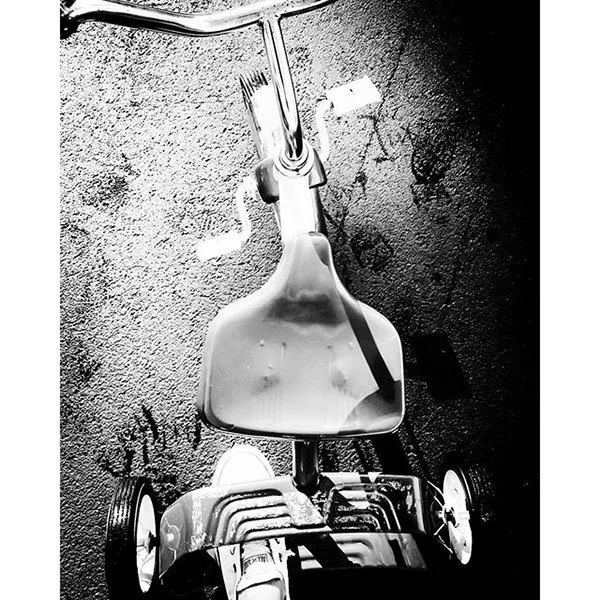 A black and white photograph of a small, four-wheeled stroller with a plastic pitcher hanging from its handle bar.