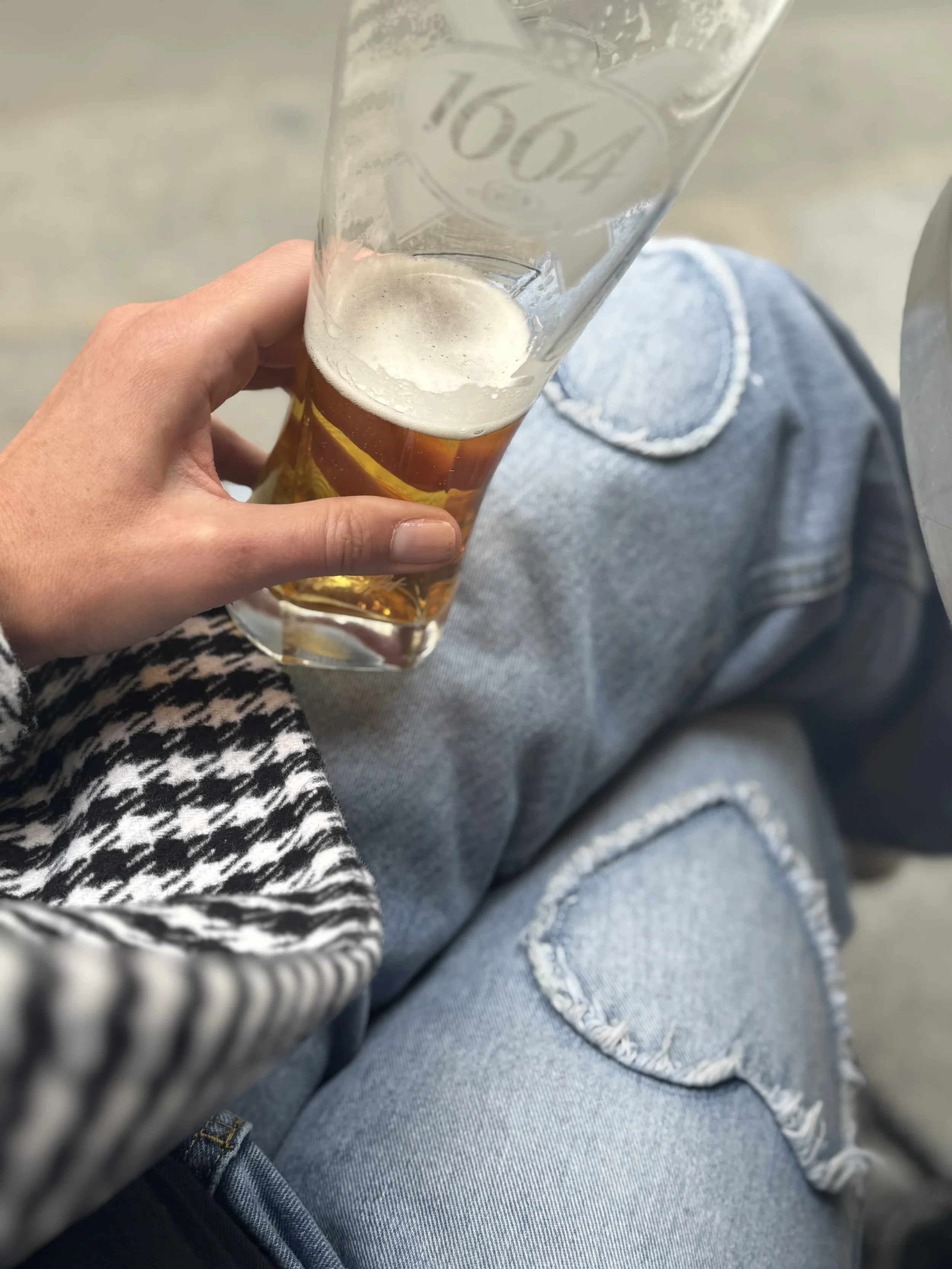 Person holding a glass of beer, wearing gray jeans and a black and white houndstooth patterned sleeve.
