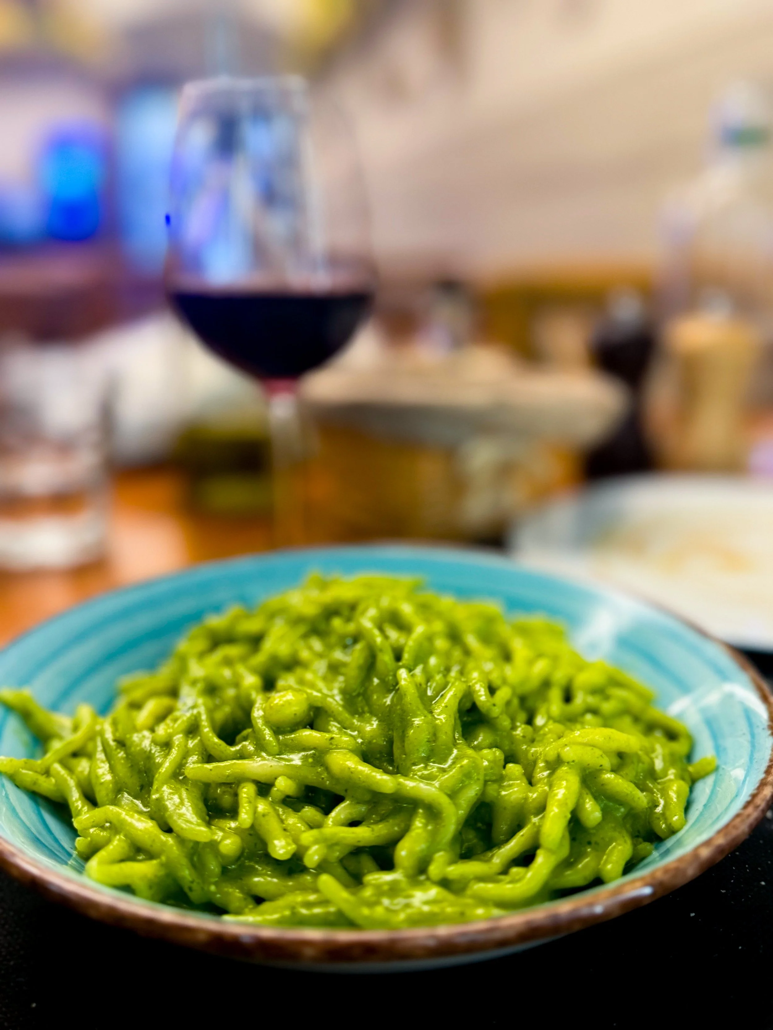 A blue bowl filled with green pasta noodles in the foreground, with a blurred background showing various bottles and kitchen items.