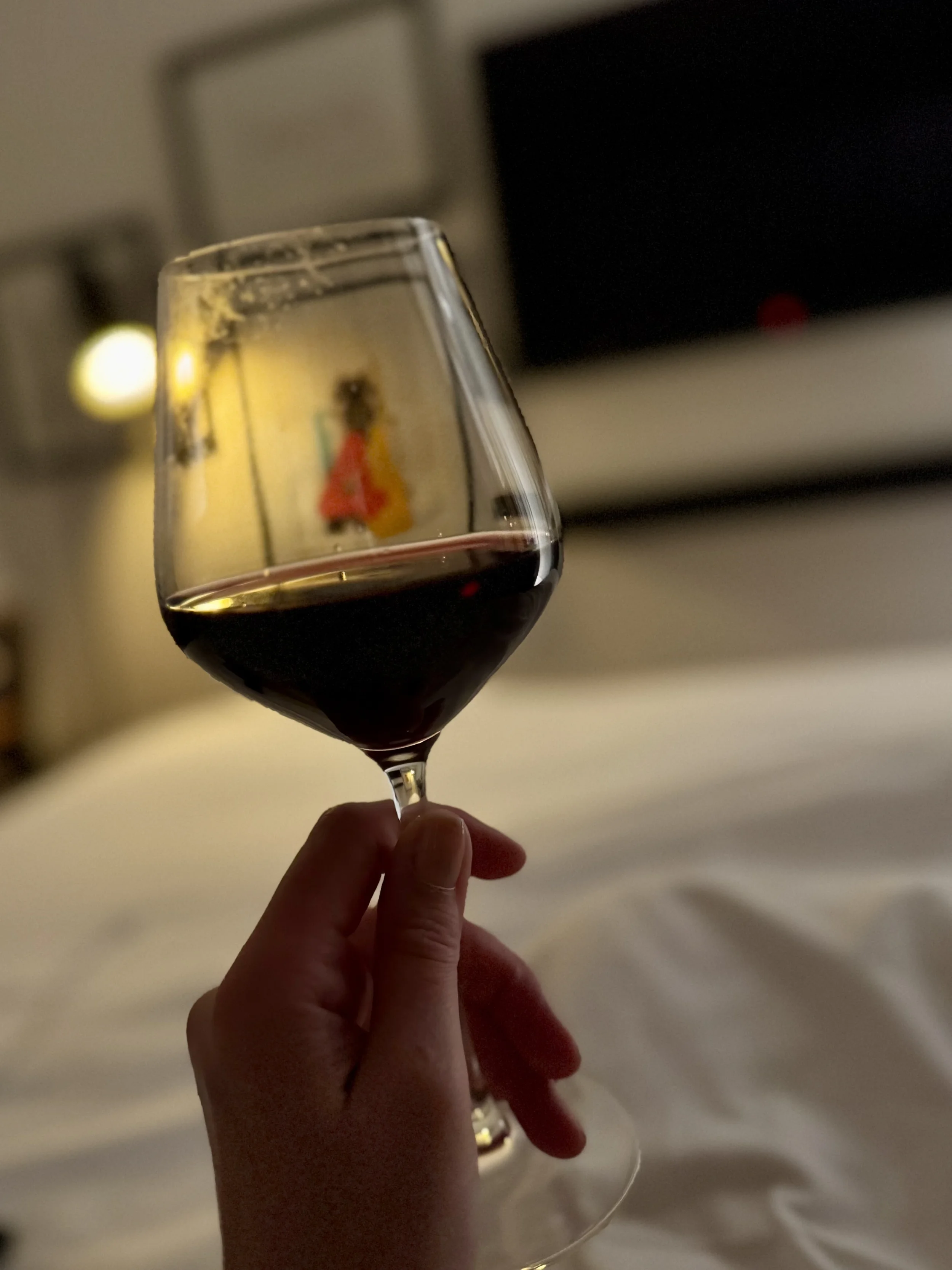 A person holding a glass of red wine in front of a blurred TV screen and wall in a dimly lit room.