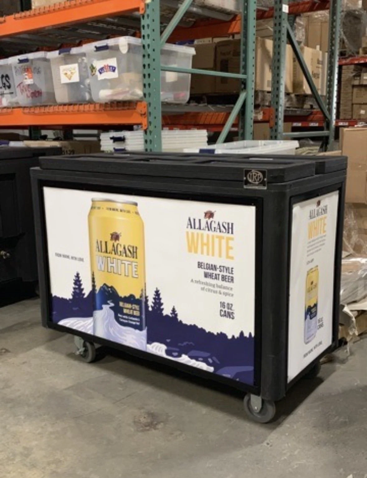 A portable display featuring Allagash White Belgian-style wheat beer, with a mountain and pine tree design on the signage, located in a warehouse or store aisle.