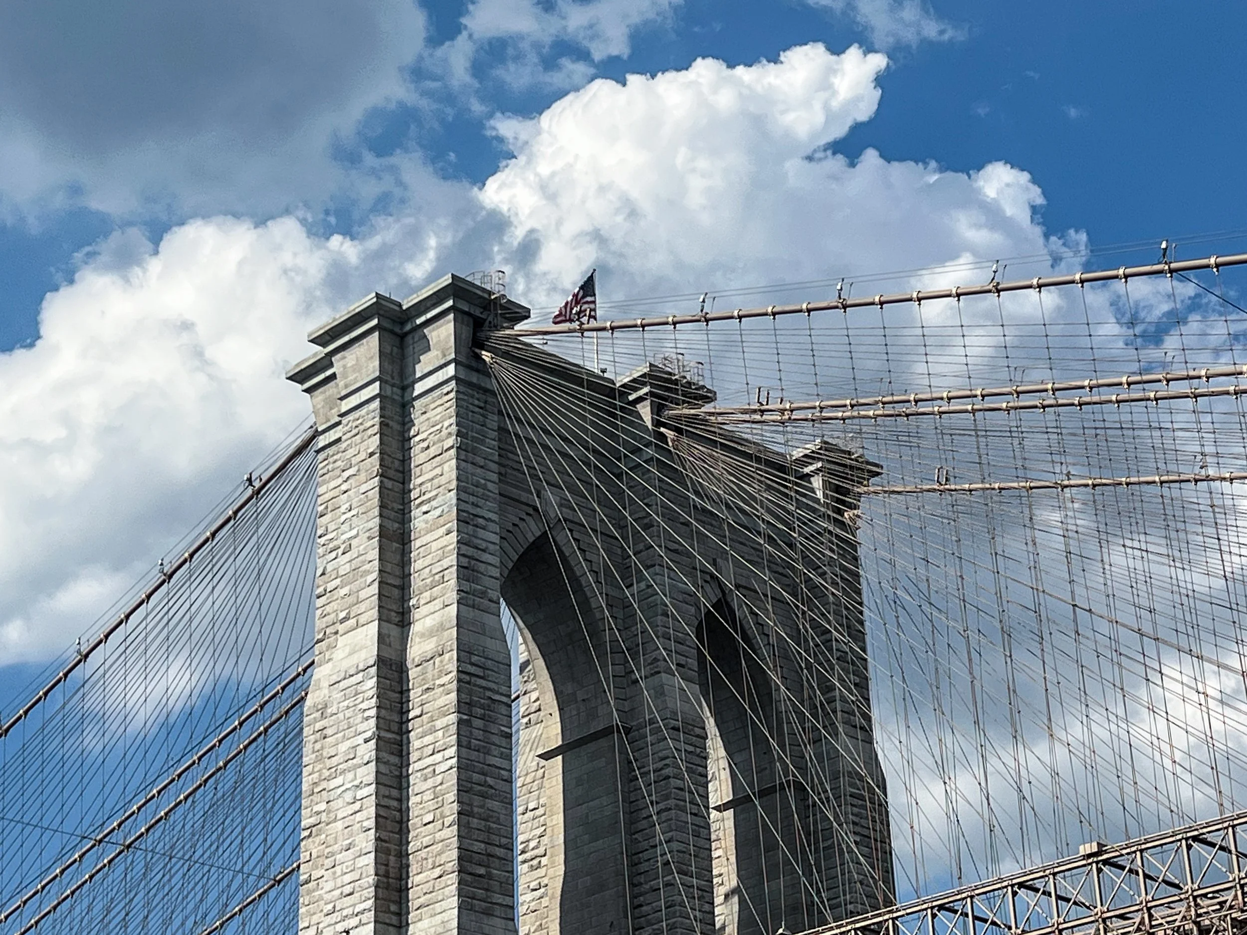Close-up of Brooklyn Bridge's stone tower with American flag, steel cables extending outward, against a background of blue sky with white clouds.