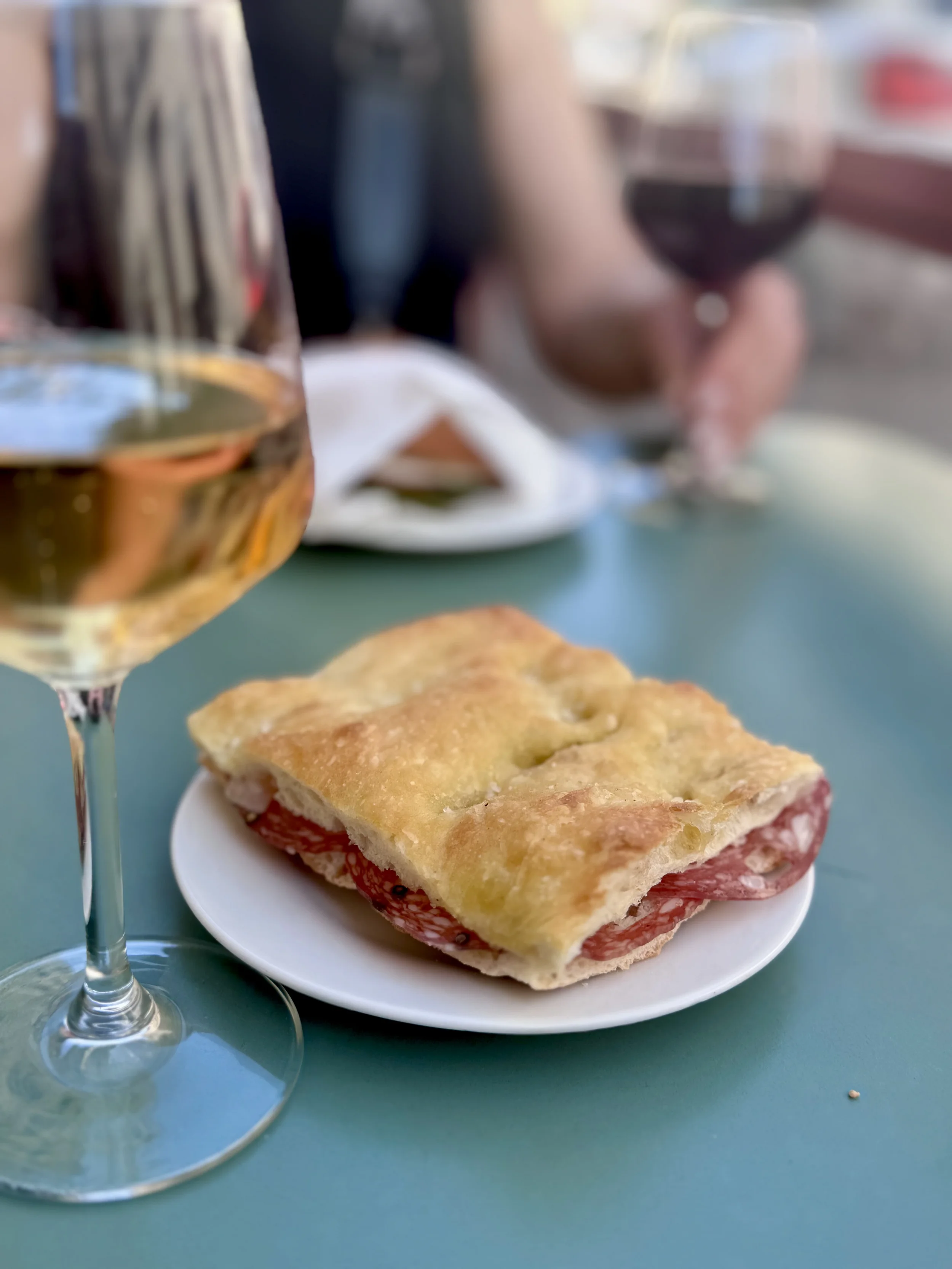 A glass of white wine, a plate with a slice of pizza topped with cheese and tomatoes, and a blurred background with a person holding a glass of red wine.