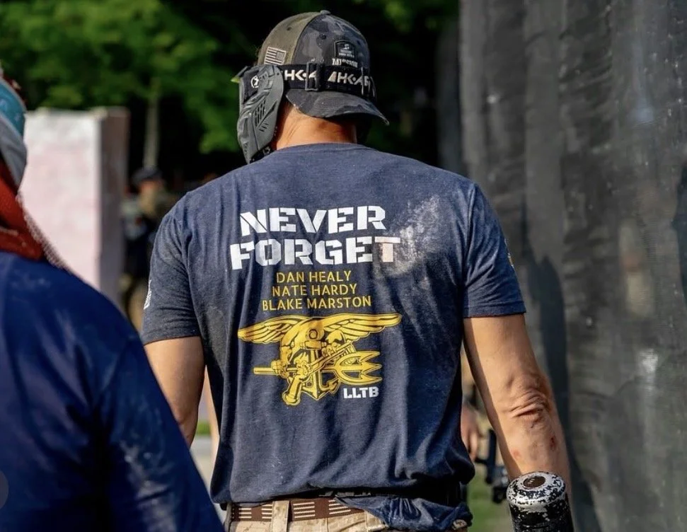 Back of a person wearing a dark blue T-shirt with military graphic and text that says "Never Forget" and names Dan Healy, Nate Hardy, and Blake Marston, at an outdoor event with other people and a black fence.