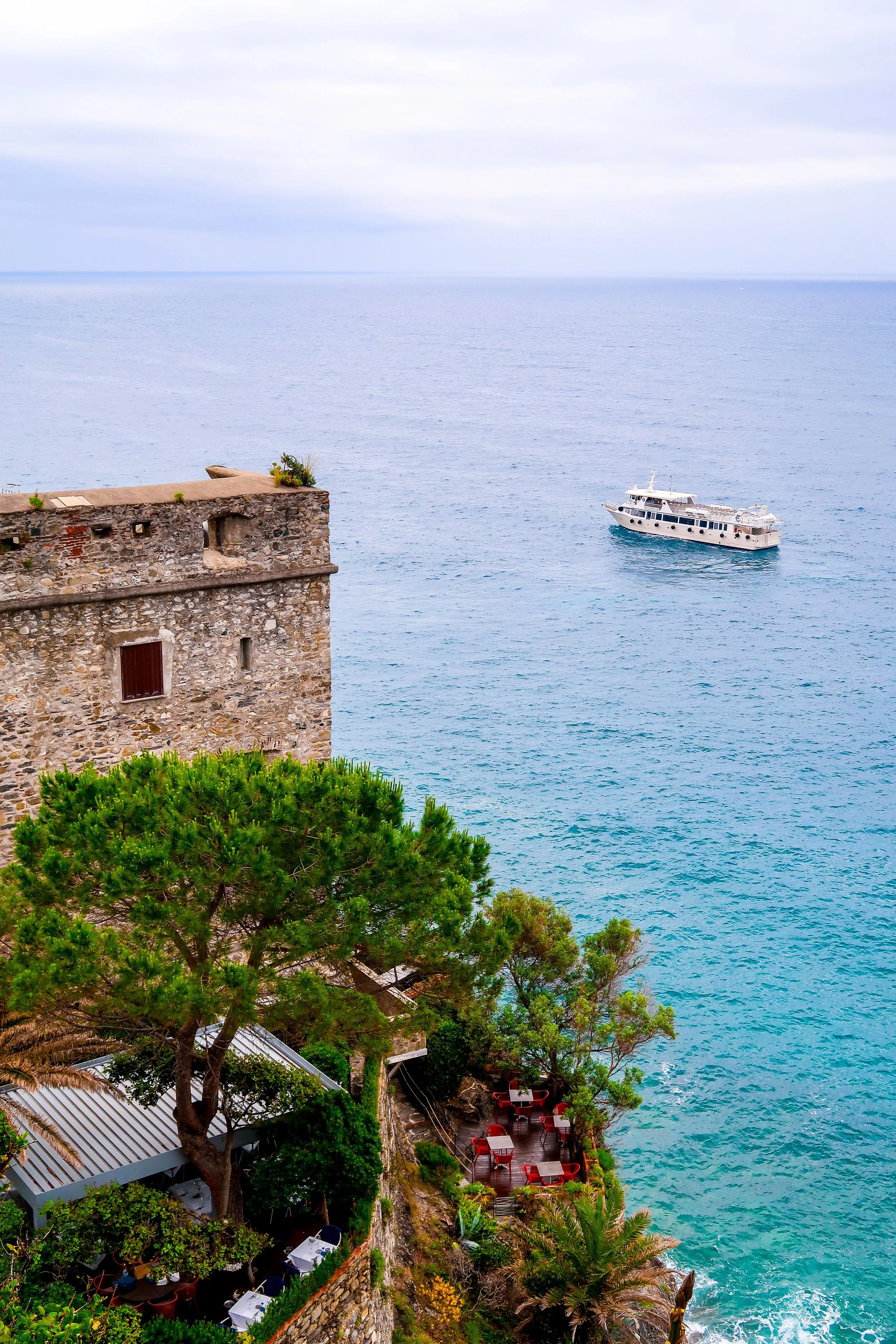 A view of a stone building with a small balcony and potted plants, overlooking the ocean. There is a tree with lush green foliage in the foreground, and a small outdoor seating area with red chairs and tables on a terrace built on the rocky coast. A 