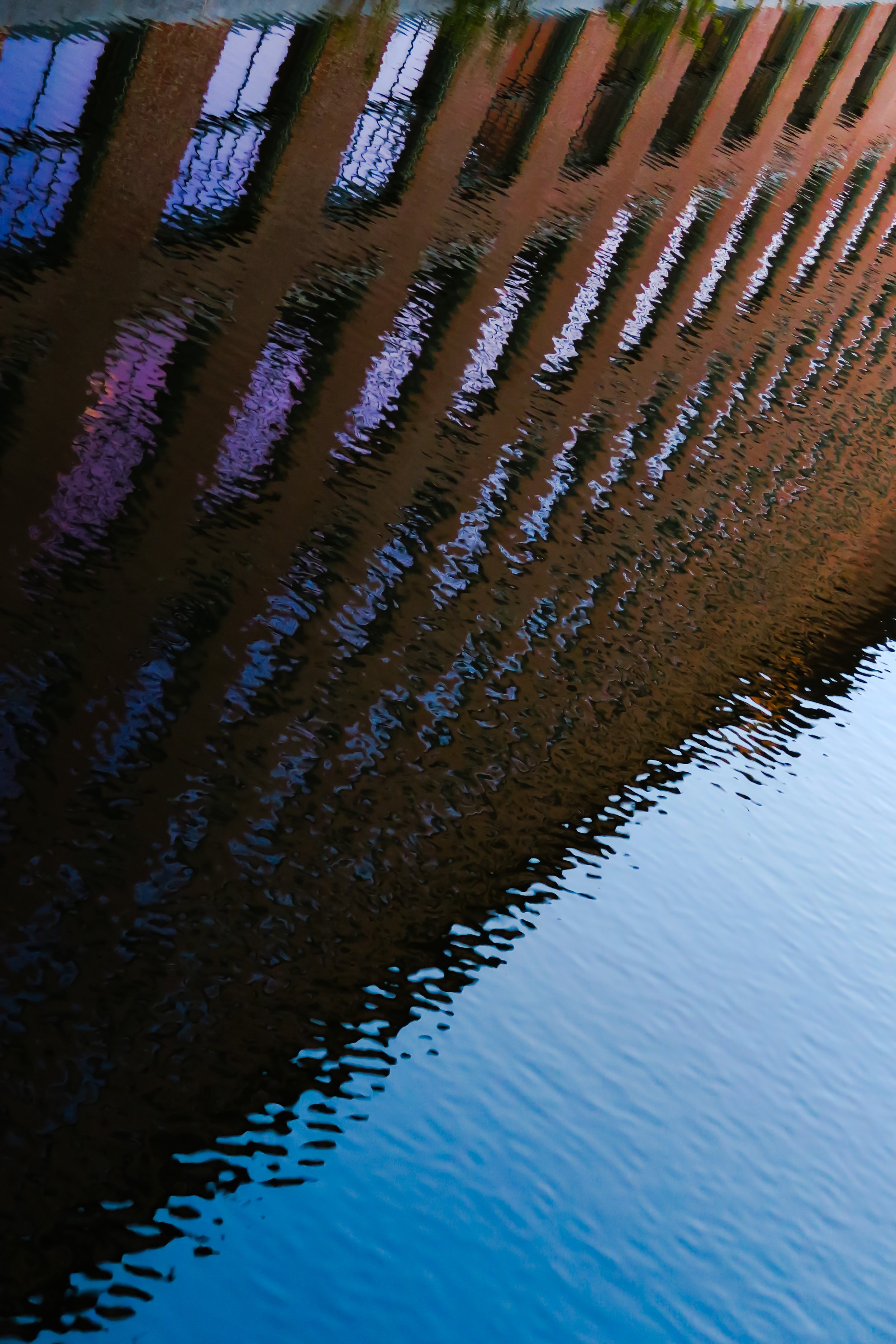 Reflected building with vertical windows and a fence on a body of water, creating a rippled mirror image.