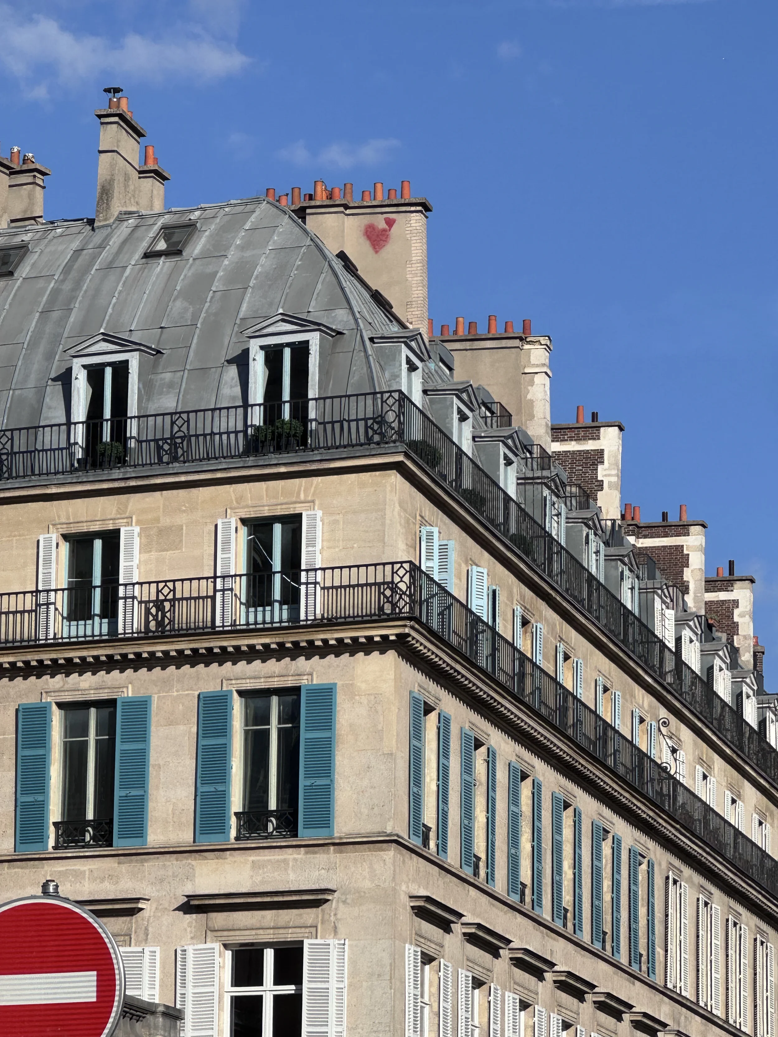 Parisian building with classic architectural style, featuring blue shutters and black wrought-iron balconies, under a clear blue sky with a small graffiti heart and letter 'Y' on chimney.