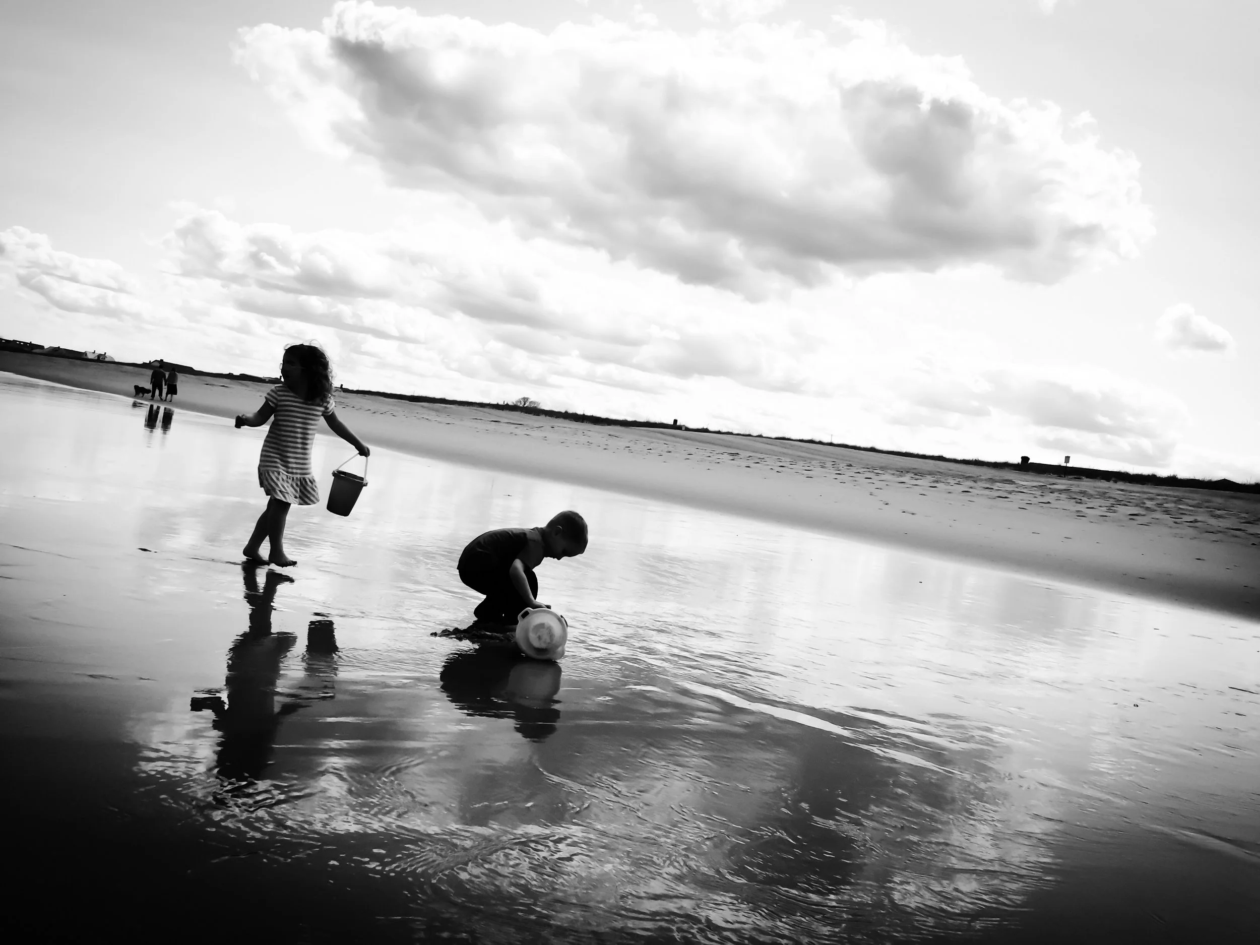 Children playing with a bucket and toy on the beach near the water, with clouds in the sky.