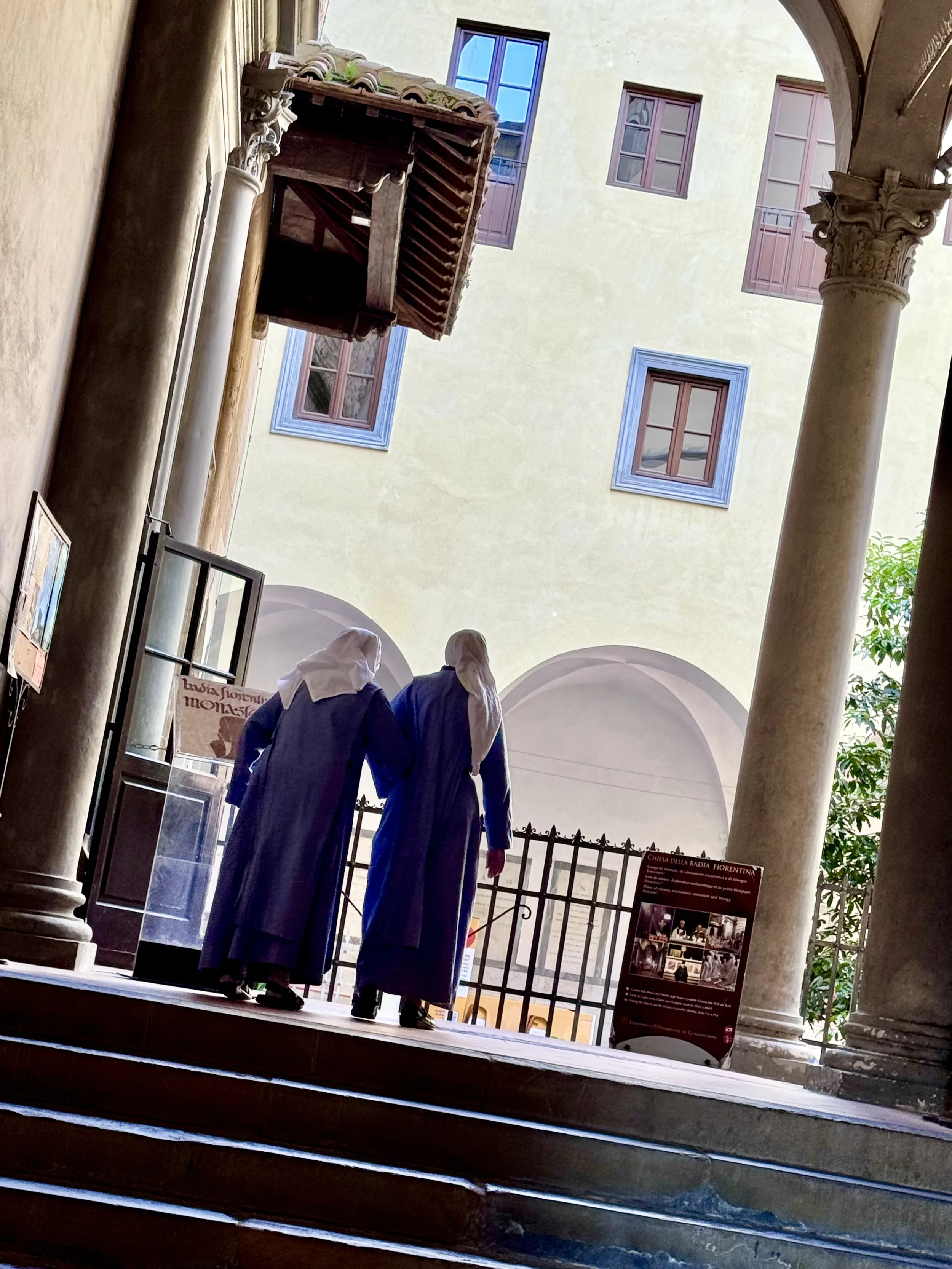 Three people dressed in historical costumes, wearing long blue dresses and white head coverings, standing on a staircase inside a historic building with columns and arches, near informational signs.