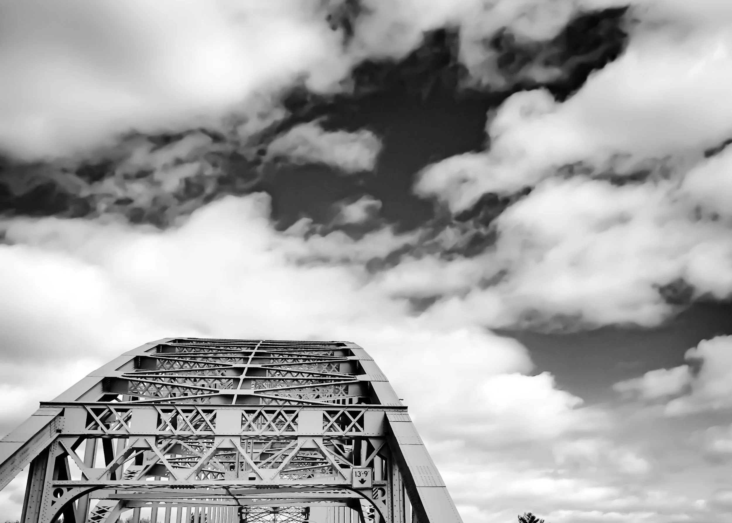 Black and white photo of a metal bridge with a geometric lattice structure, viewed from below, with a cloudy sky in the background.