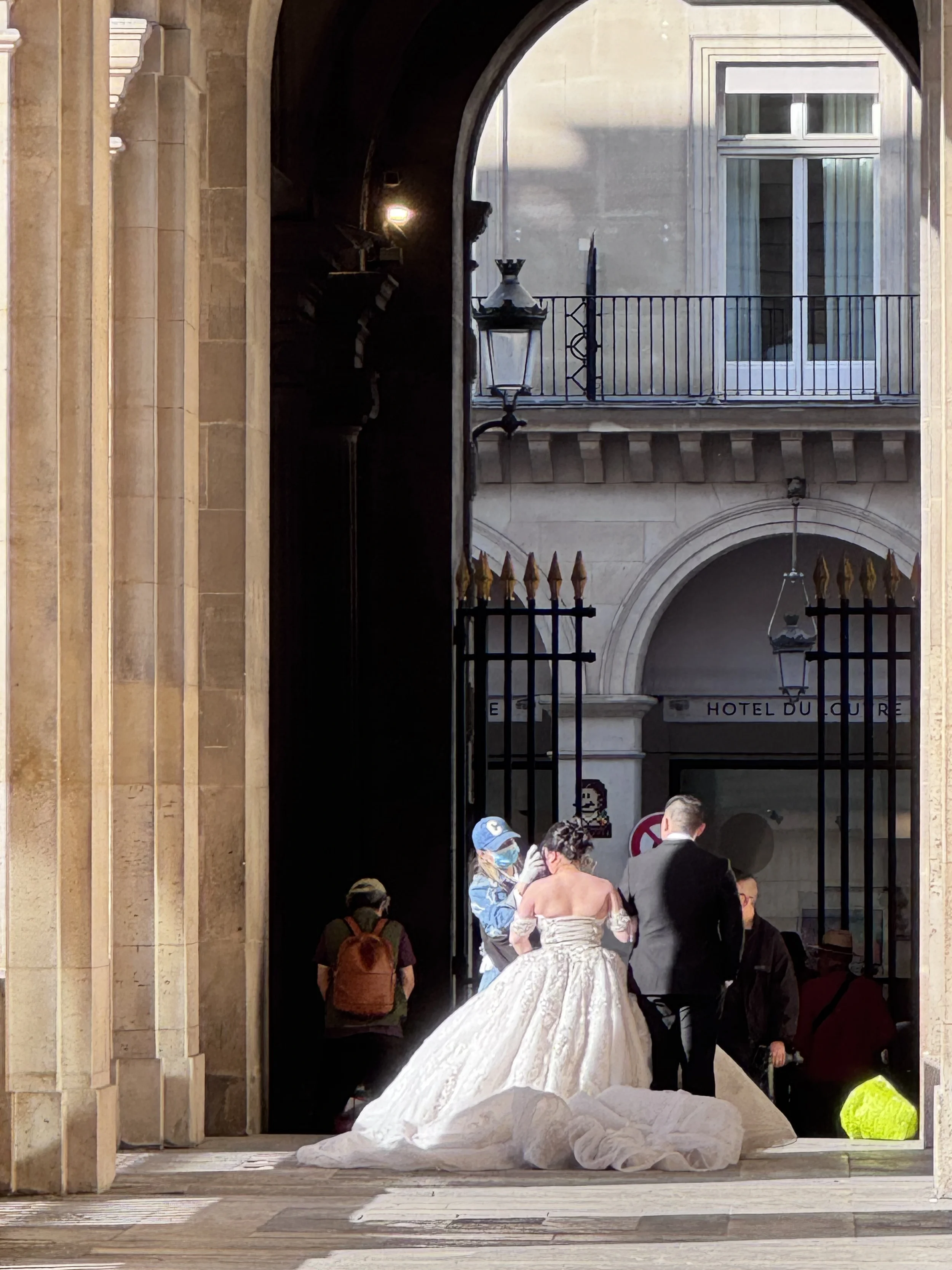A bride in a white wedding dress and veil walking through a stone archway, surrounded by people, with a large wedding dress train trailing behind her.