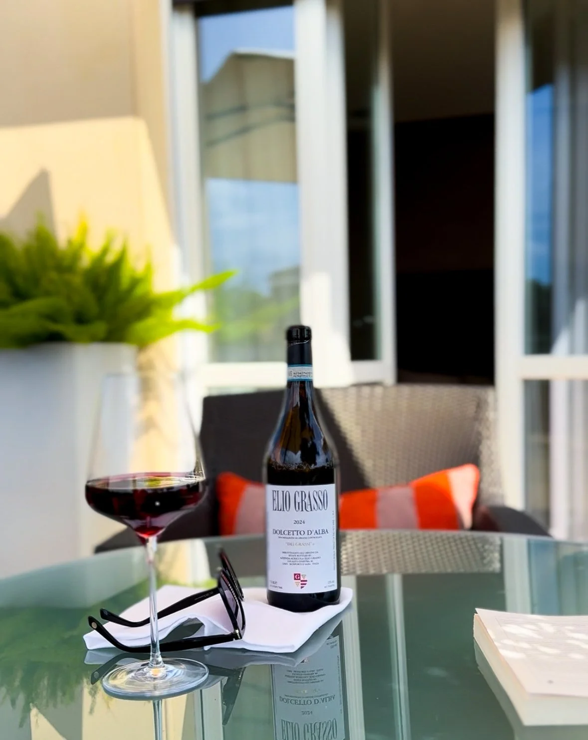 A glass of red wine, a bottle of wine labeled Elio Grasso Dolcetto D'Alba, a pair of black sunglasses, and a notebook are on a glass table outside, with a patio and a glass door in the background.