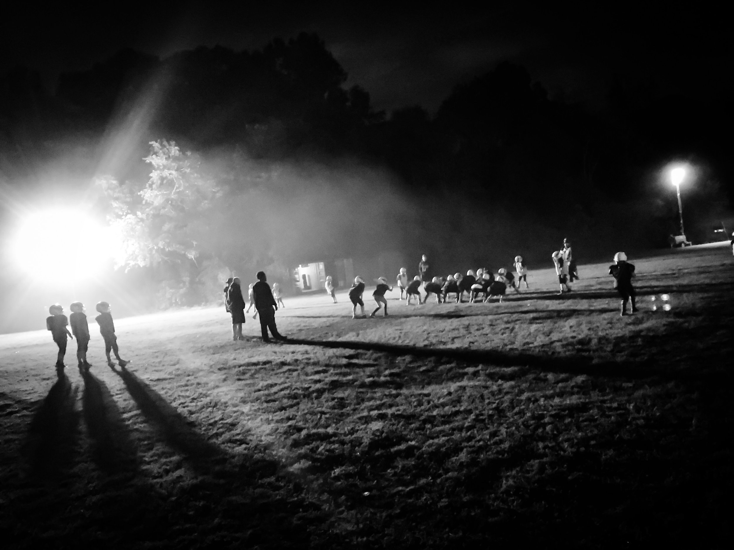 Nighttime scene of children playing football on a grassy field, illuminated by two bright streetlights, with trees and a dark sky in the background.