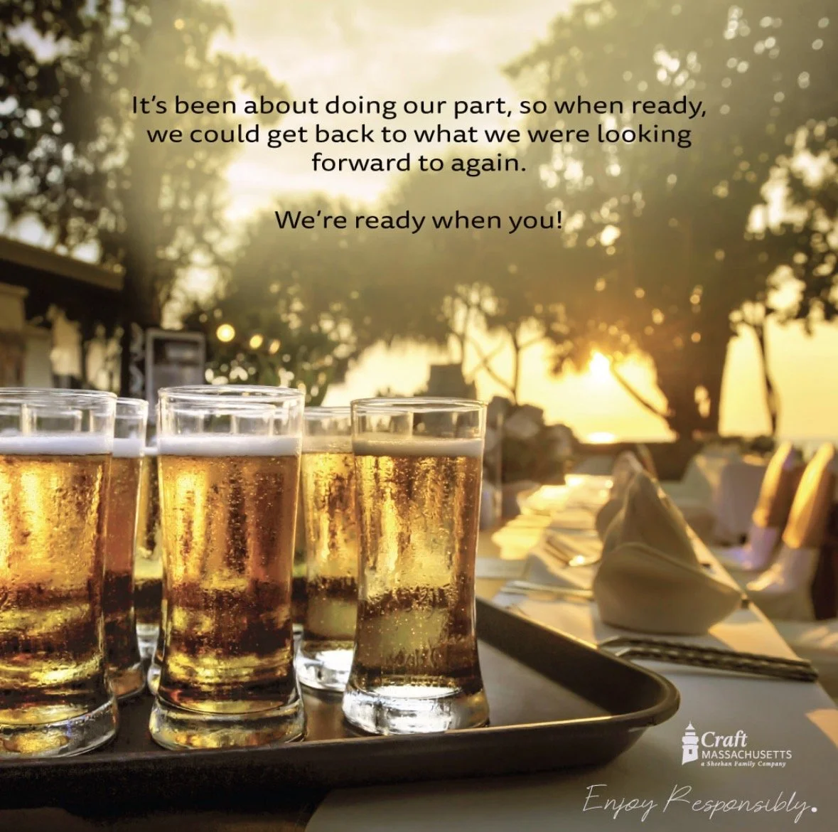 A tray with six glasses of beer on a table, set outdoors during sunset with trees and a string of lights in the background, and a dining setup with napkins and silverware.