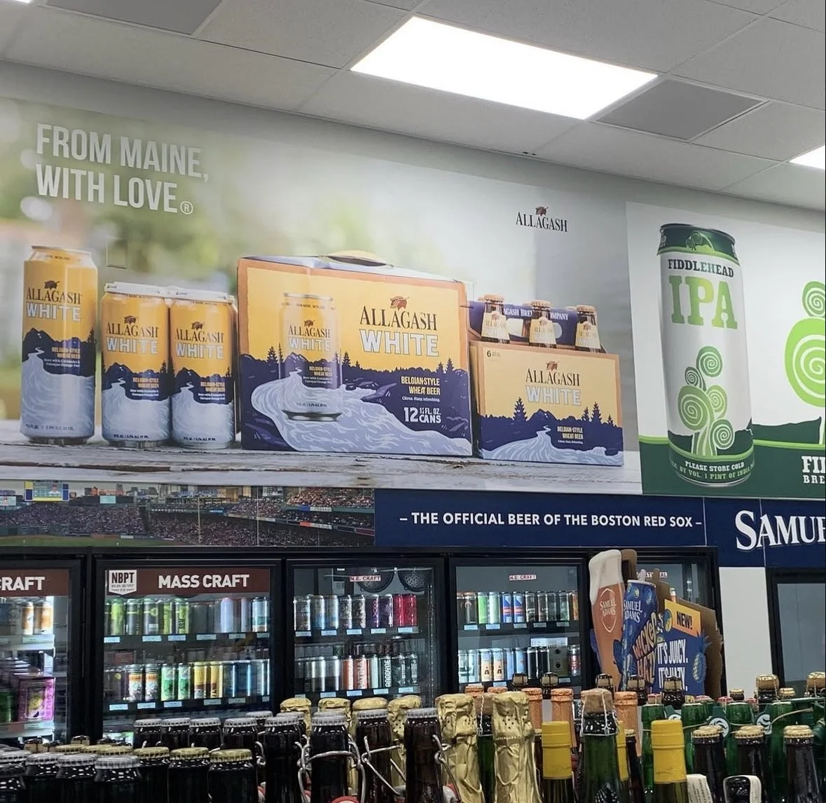 Display shelf with cans of Allagash White beer, boxes of six-packs, and refrigerators with various beverages in a store. Promotional posters for Allagash White and Fiddlehead IPA are on the wall above.