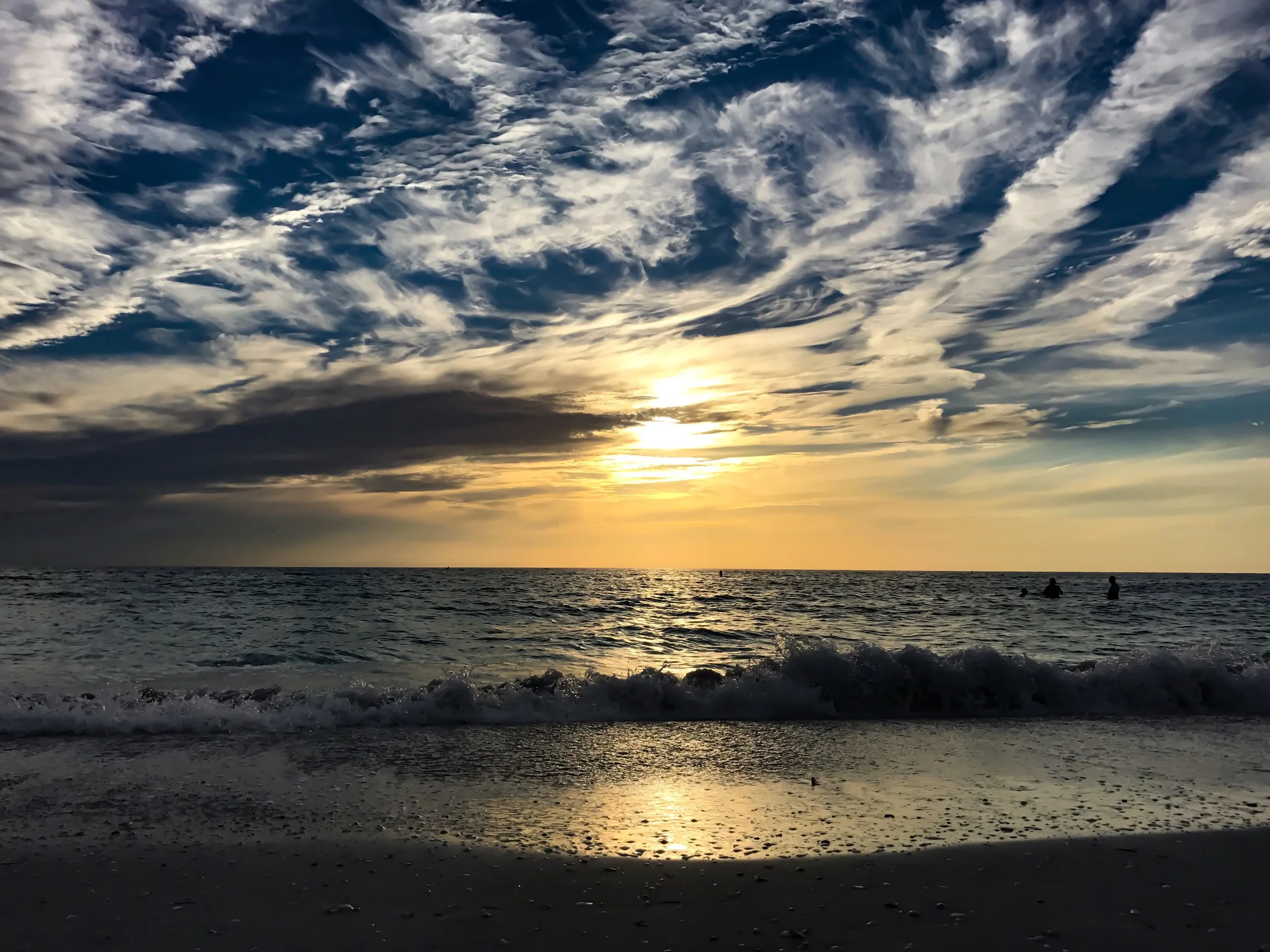 Sunset over the ocean with a sky filled with scattered clouds, and silhouettes of people in the water, with waves gently crashing on the shore.