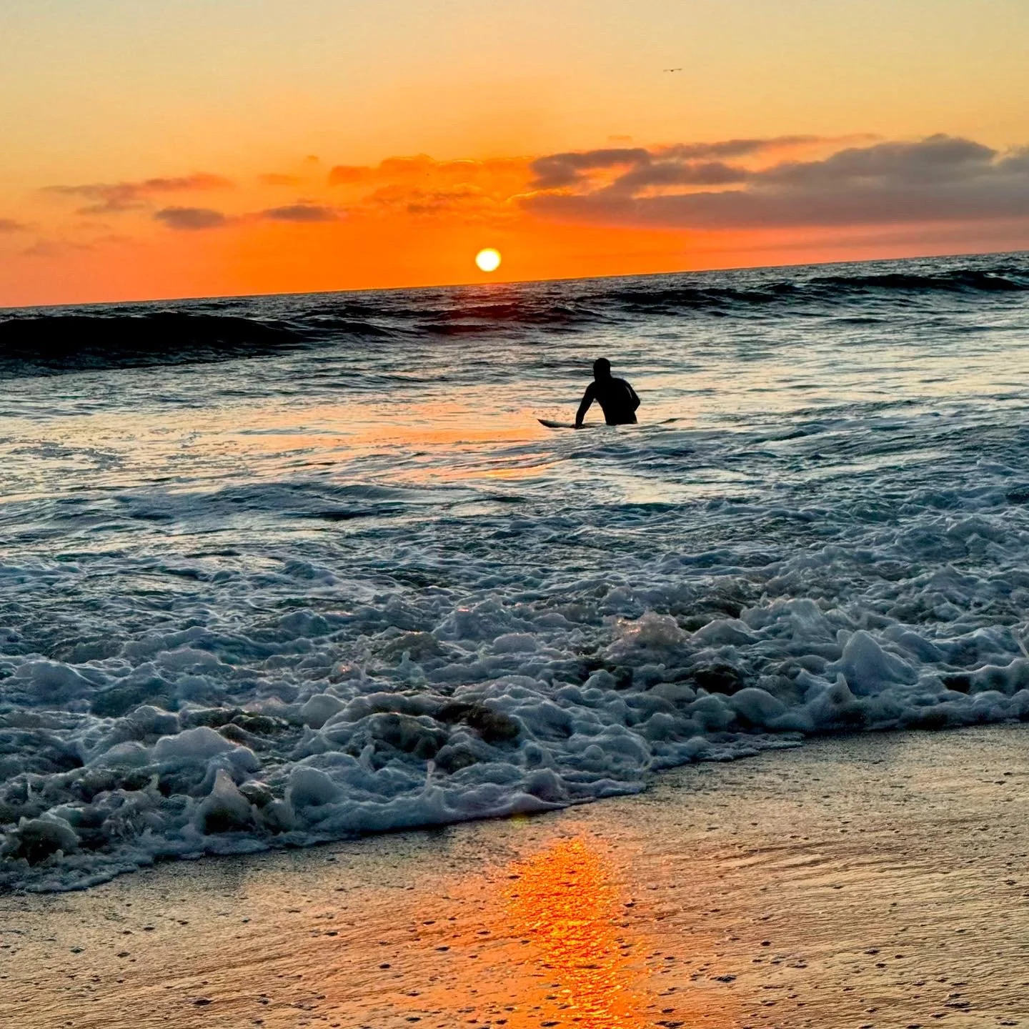 A person on a surfboard in the ocean during sunset, with waves and foam in the foreground and the sun setting on the horizon.