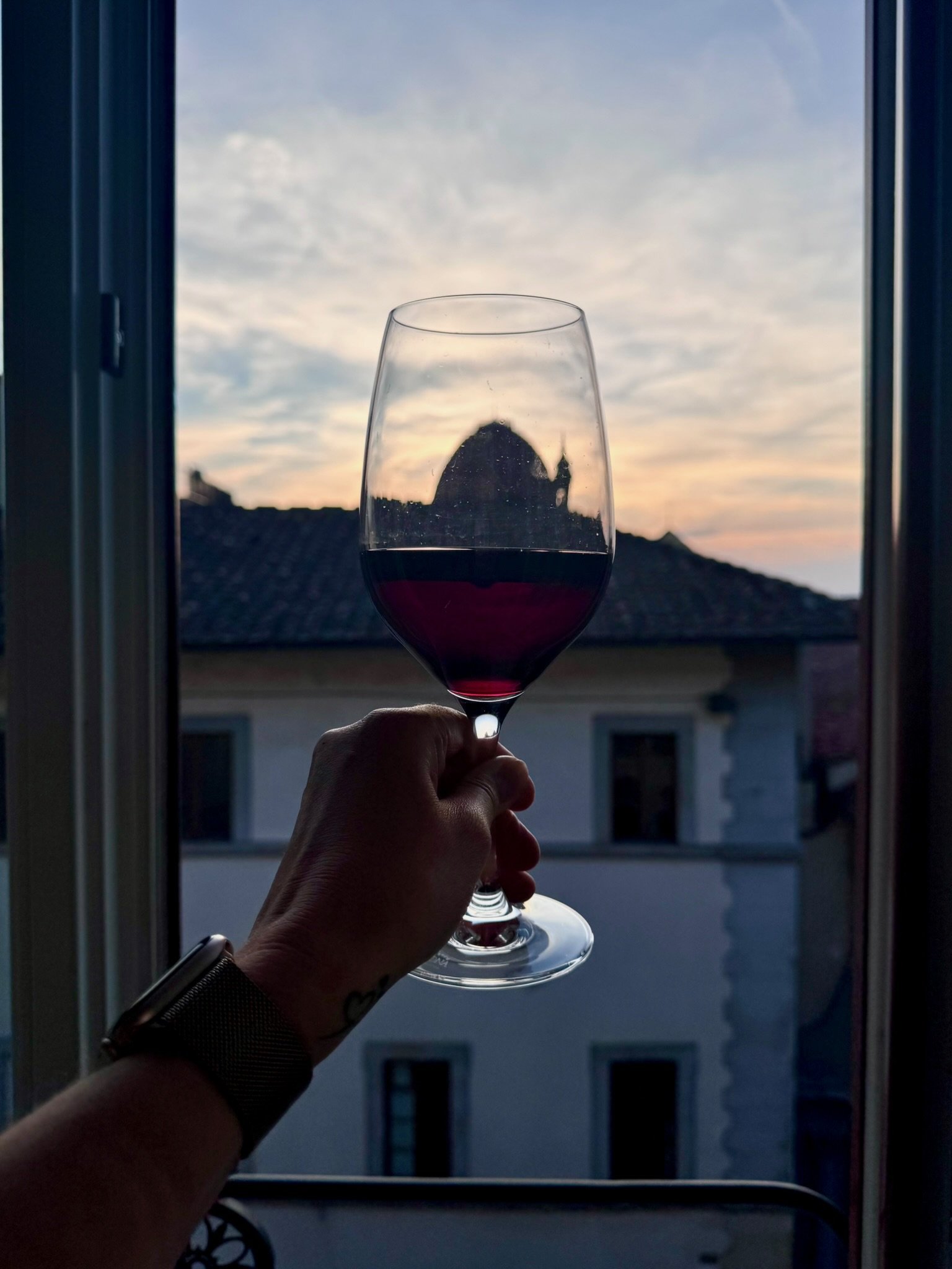 A hand holding a glass of red wine in front of a window at sunset, with a cityscape and a prominent building in the background.