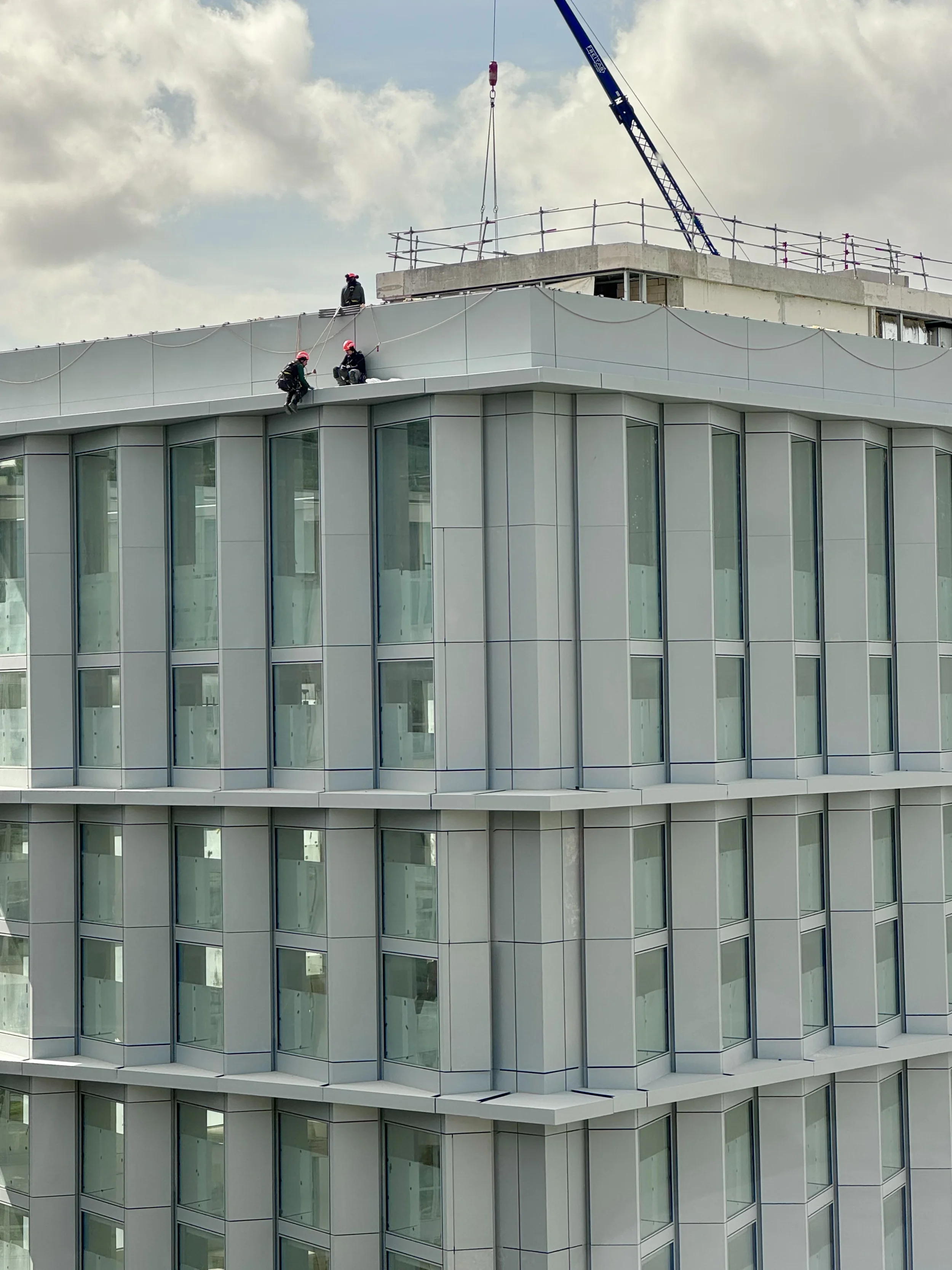 Construction workers wearing safety gear working on the top floor of a modern, glass-fronted building under a cloudy sky, with a crane nearby.