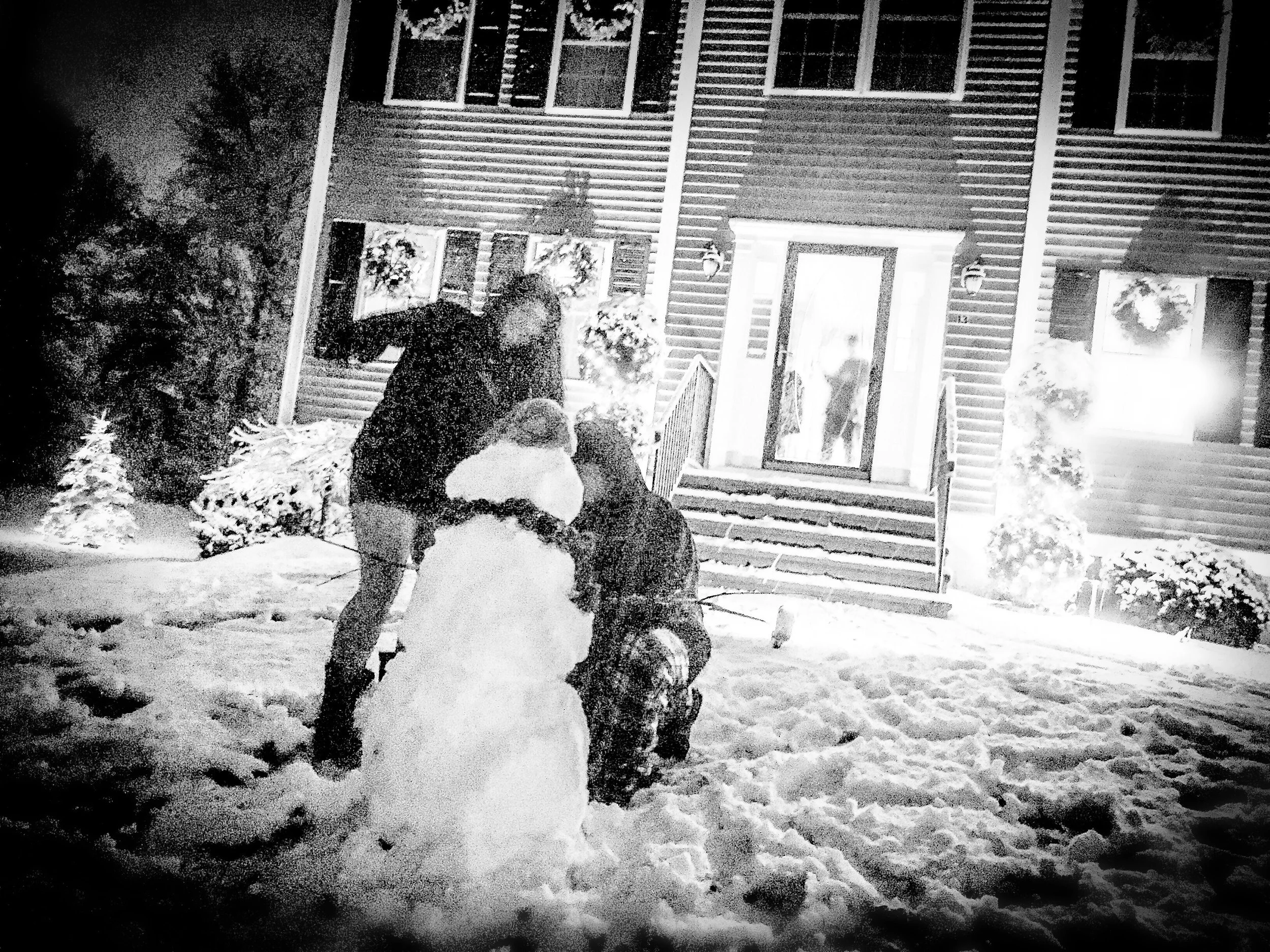 A black and white photo shows people walking in a snowy yard in front of a house decorated with holiday wreaths and lights, with snow-covered bushes and trees around.