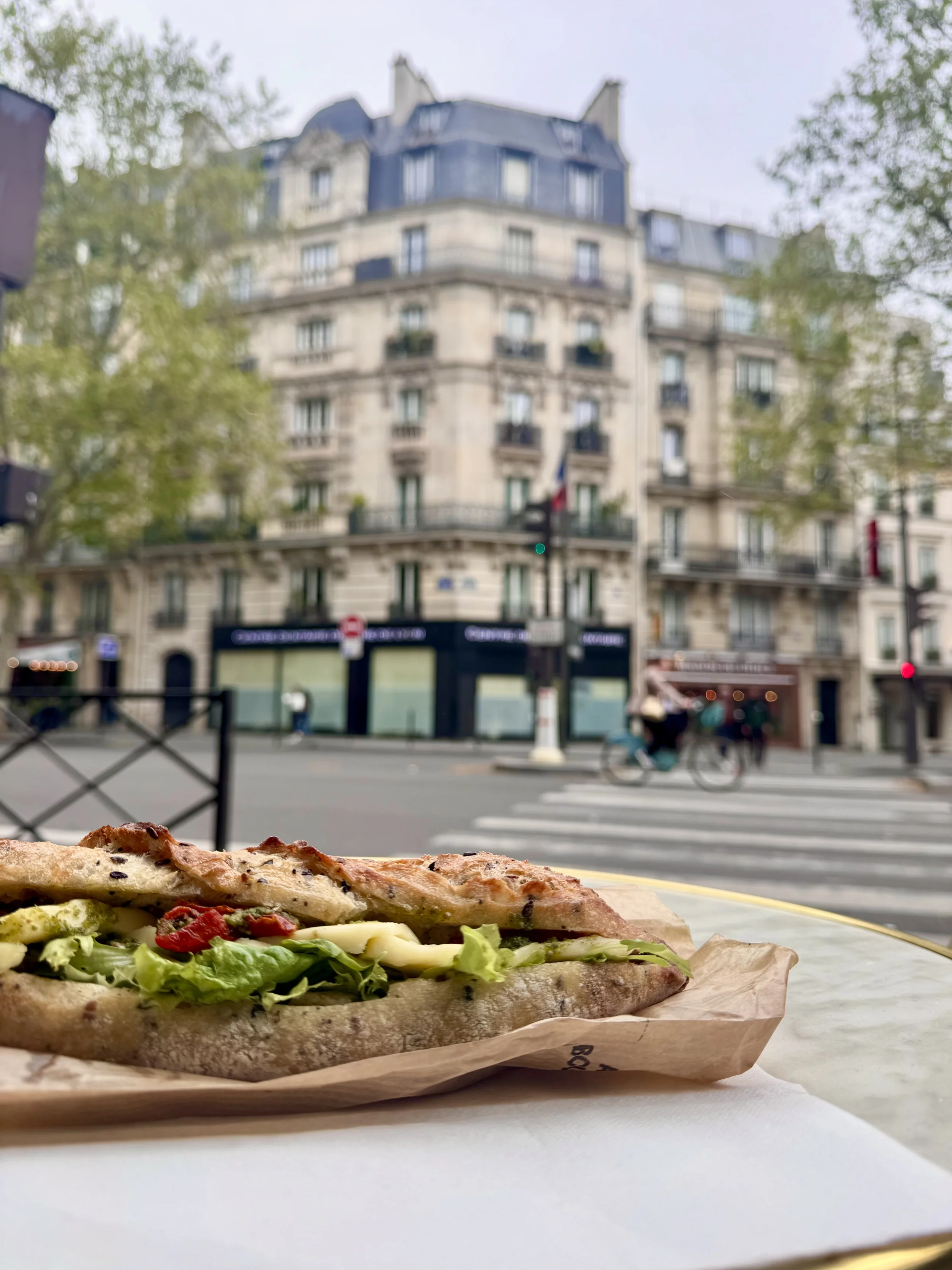 Close-up of a pizza with various toppings on a paper tray, in front of a blurred city street scene with a large building and pedestrians.