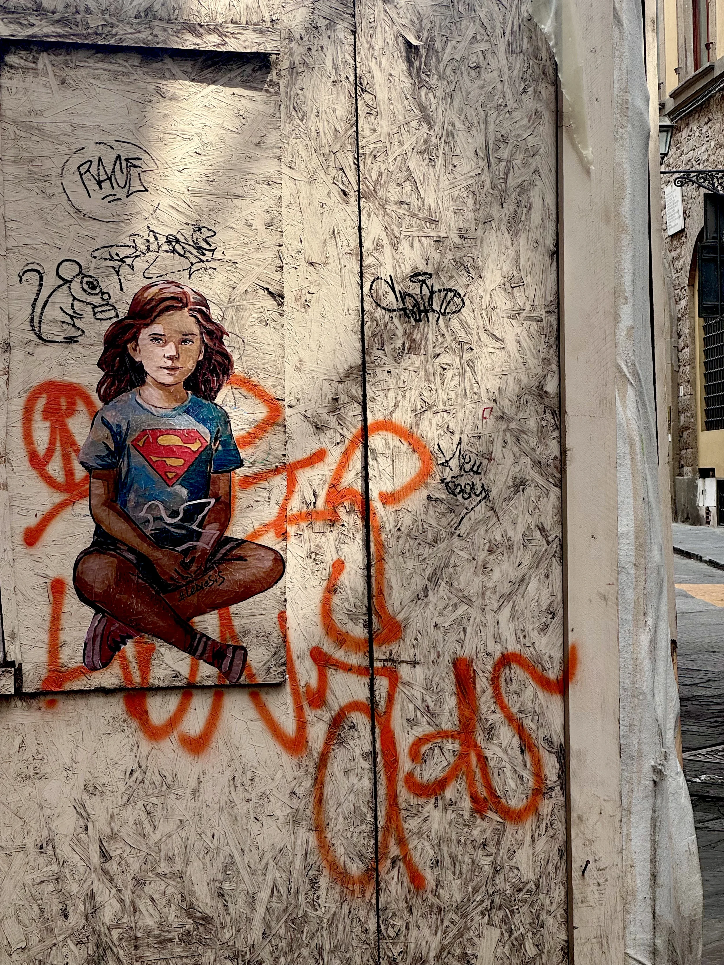 Graffiti art of a young girl with long hair wearing a Superman t-shirt, sitting cross-legged with a serious expression, painted on a wooden wall with orange spray-painted tags and black graffiti.