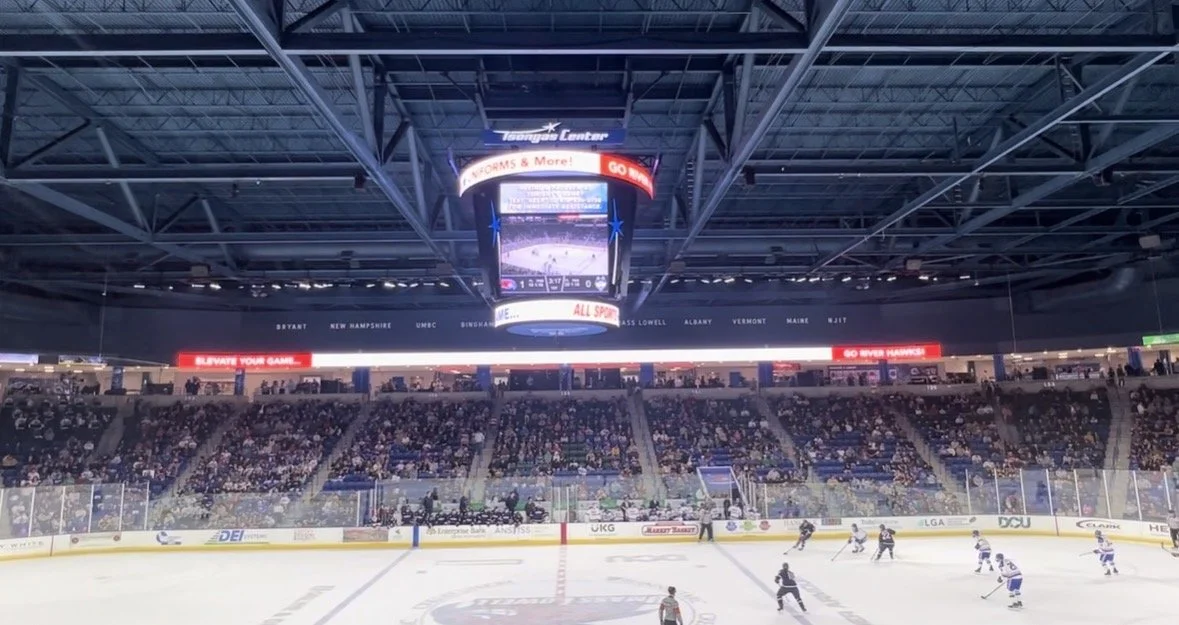 An indoor ice hockey arena with spectators seated in the stands, a game in progress on the ice, and a large digital scoreboard hanging above the rink.