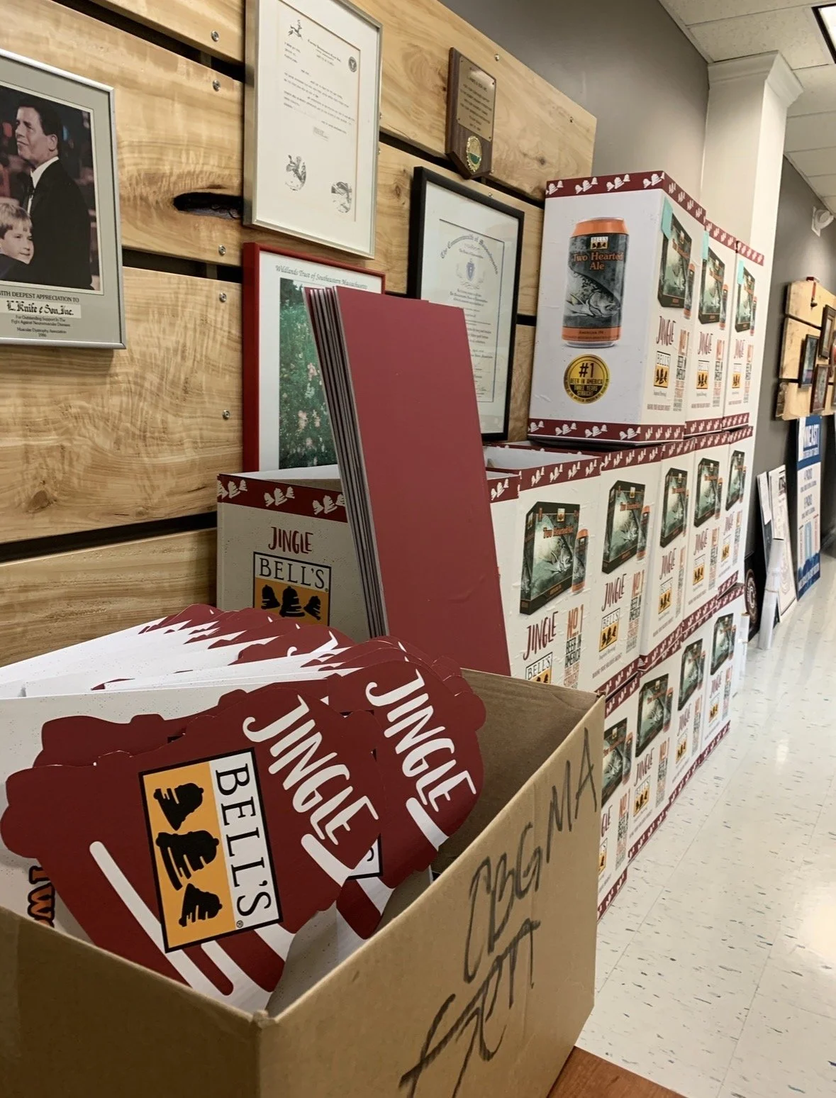 Box of Bell's Jingle Bells holiday-themed door hangers, with more boxes and framed certificates on the wall behind.