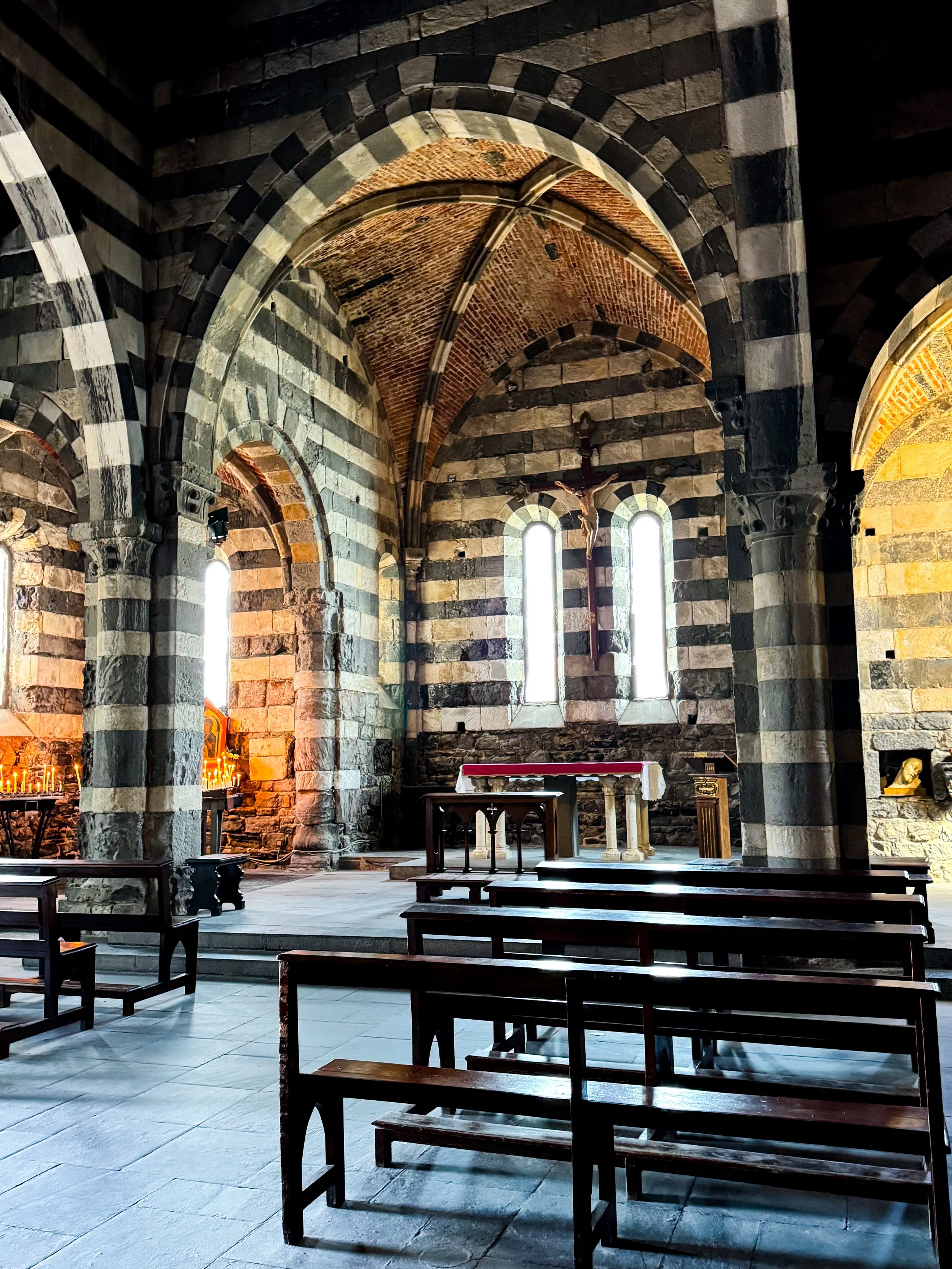 Interior of an old church with stone walls, arched ceilings, wooden benches, an altar, a crucifix on the wall, and candle lights.