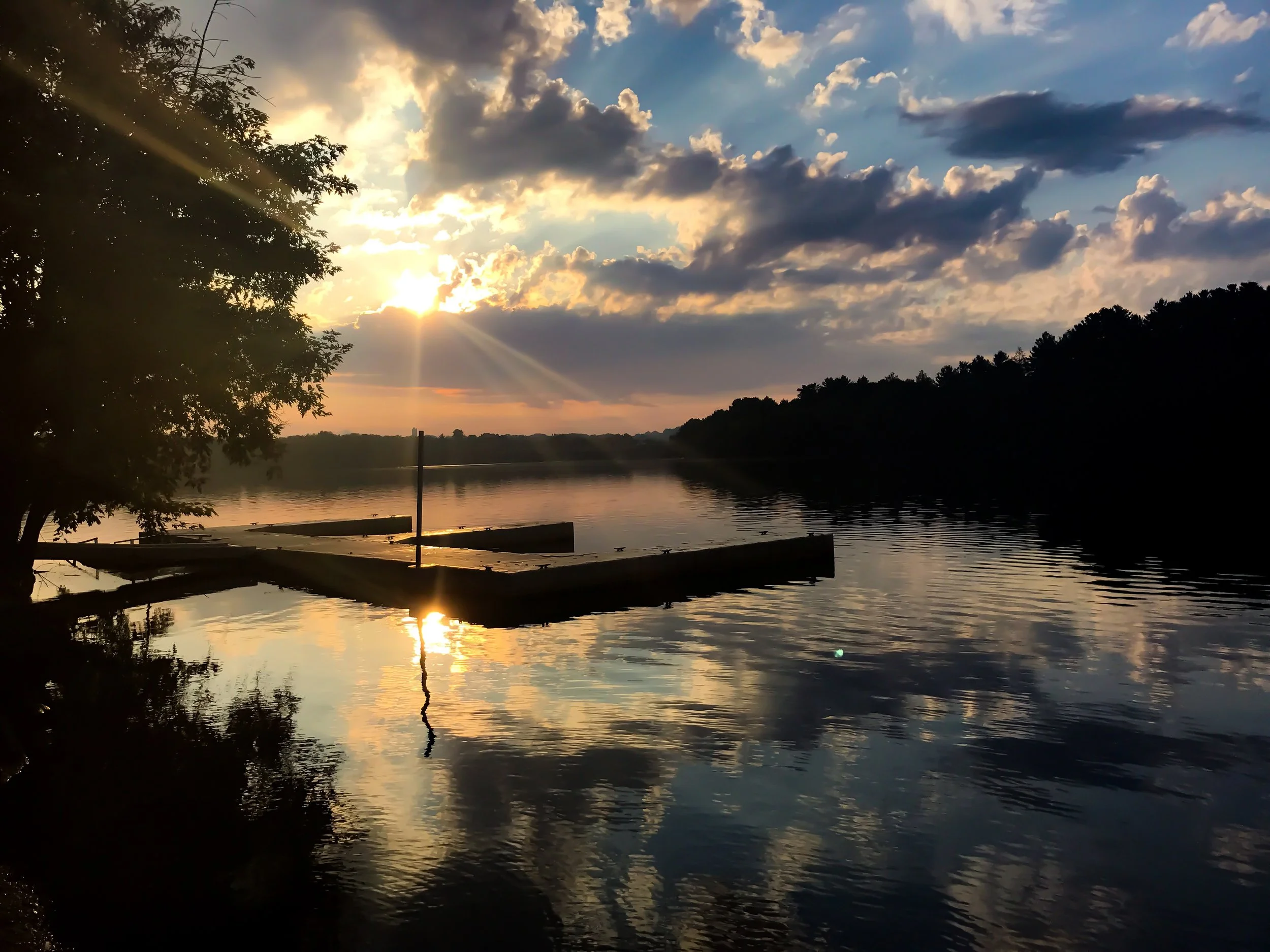 Sunset over a river with clouds in the sky, trees silhouetted on the left, and their reflection in the water.