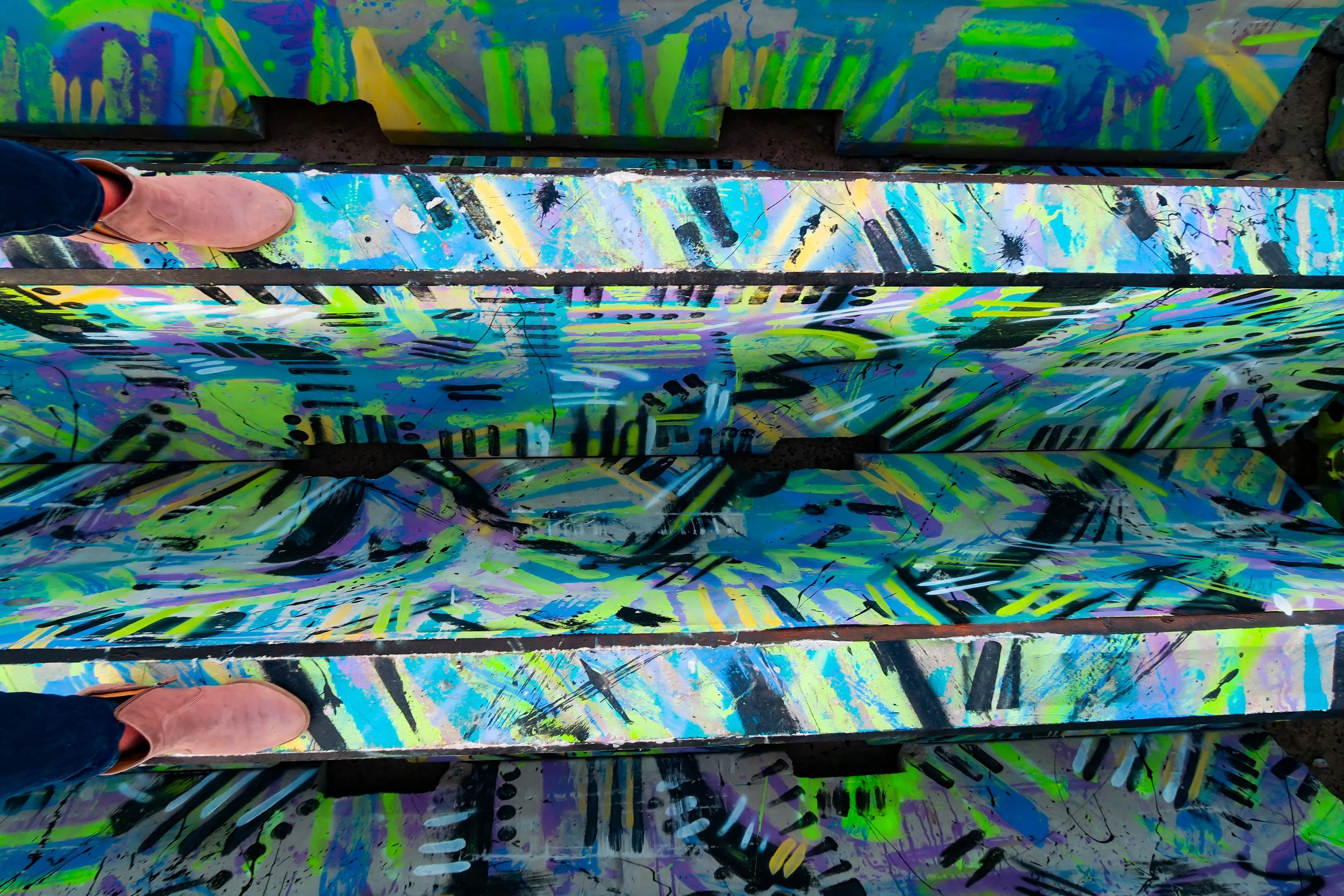 A person's foot in a pink shoe standing on a bright, colorful, abstract painted surface with black lines and splashes of neon colors.