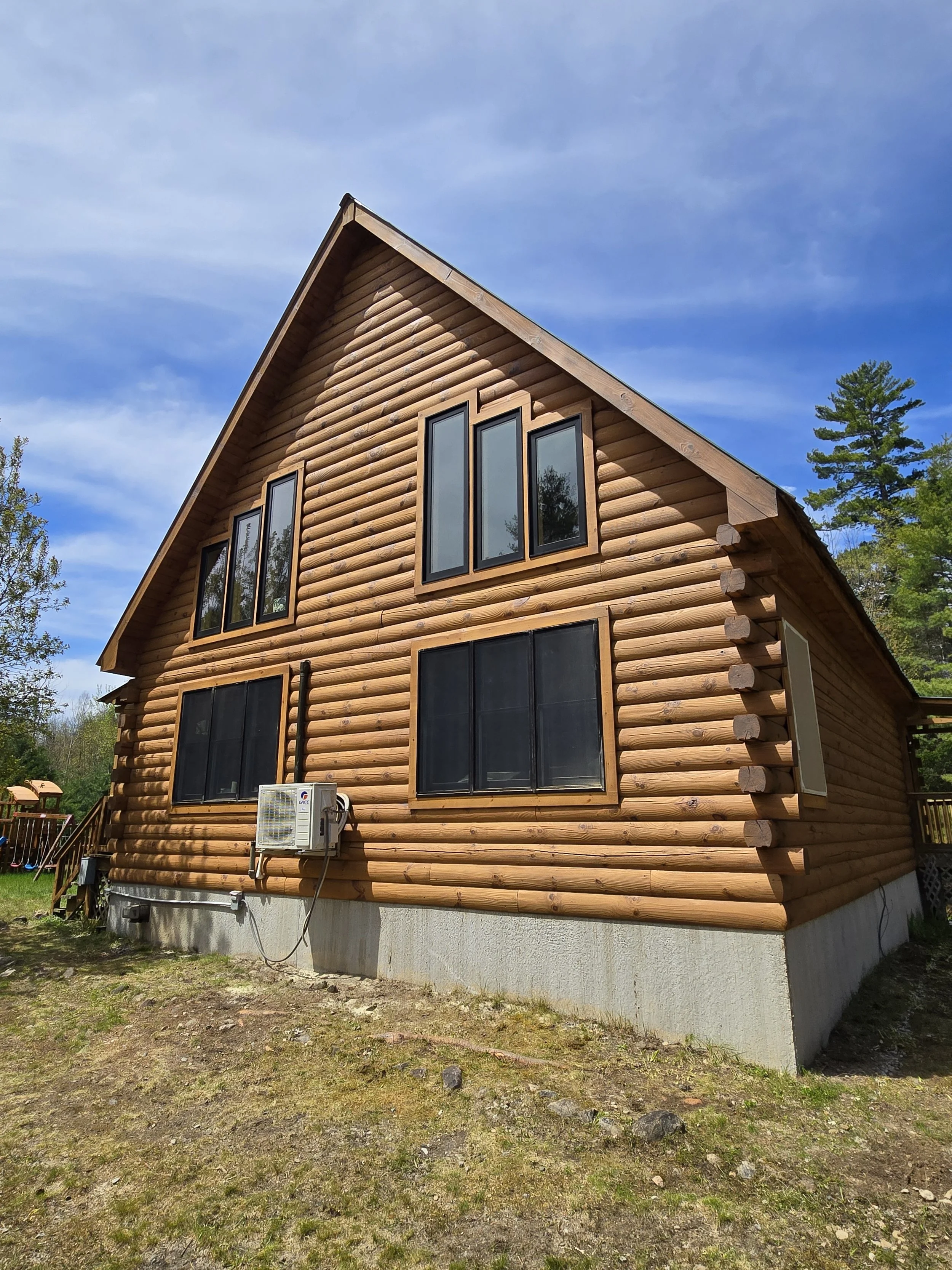 The image shows the side view of a wooden house with a steep gable roof, multiple large windows, an air conditioning unit, and a concrete foundation. There is a grassy and rocky yard and some trees in the background under a blue sky with a few clouds