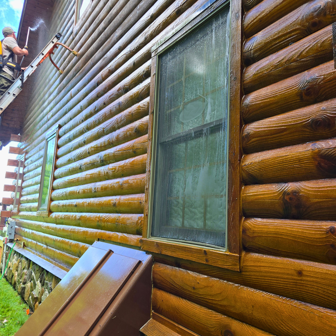A person on a ladder cleaning a window on the side of a wooden log cabin.