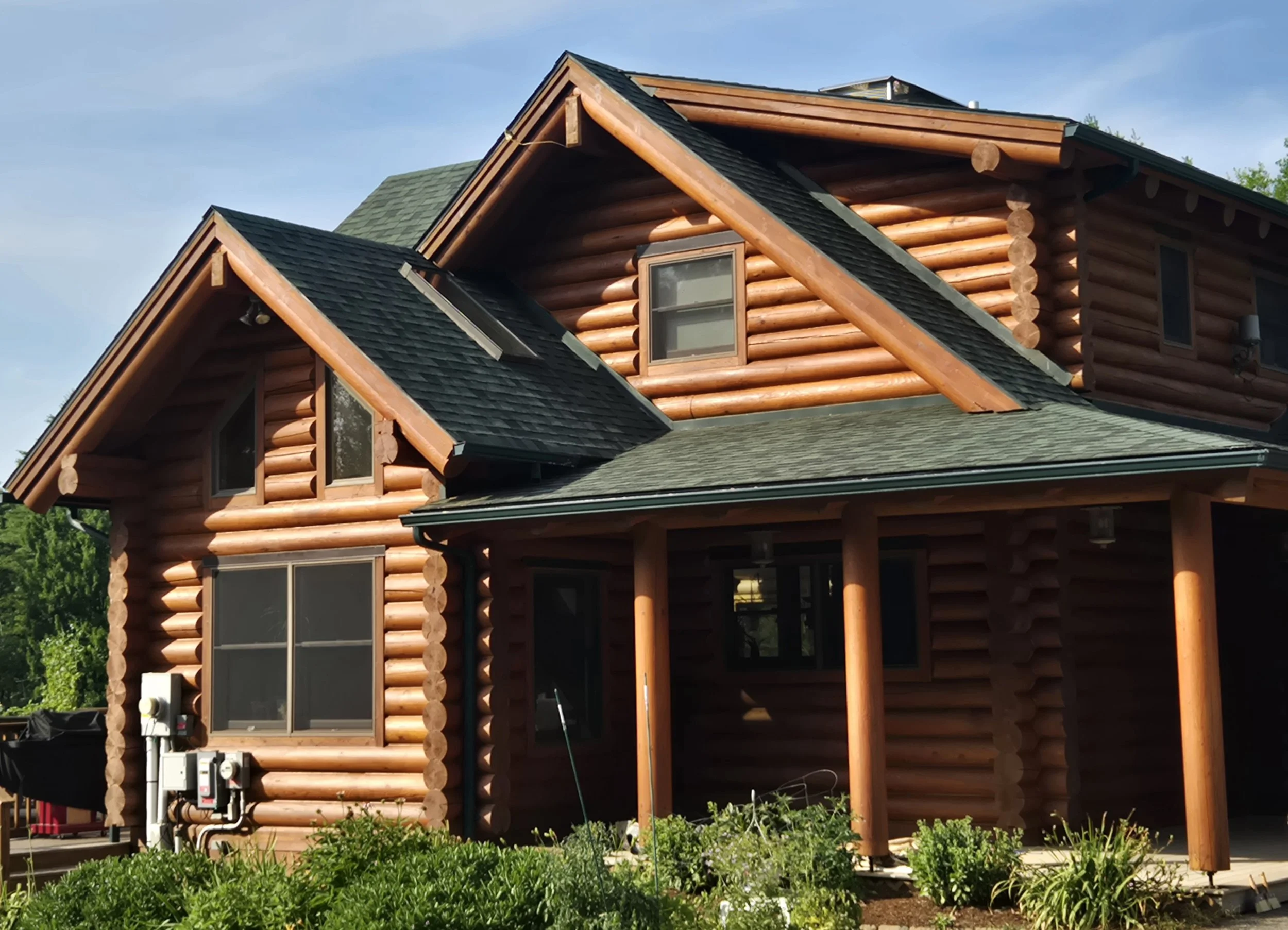 A log cabin house with a green shingles roof, multiple windows, and a wood porch overgrown with green plants under a blue sky.